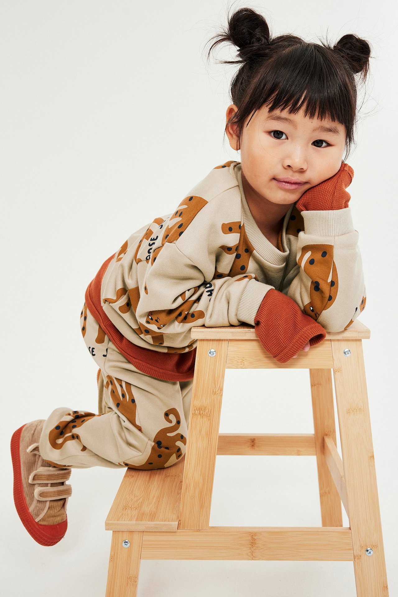 Barn i beige mønstret genser med print, mansjetter ved hals, ermer og bunning, på trebænk.