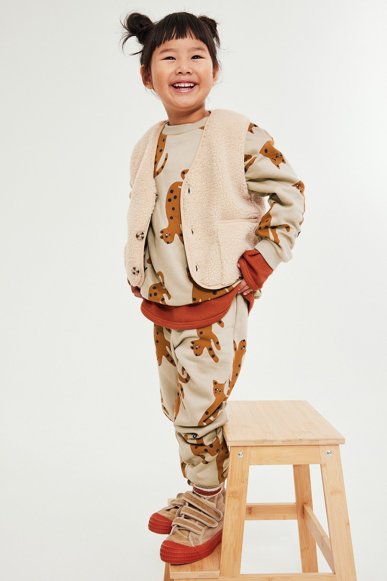 Lasten beige-kuvioitu collegepaita, fleeceliivi, beige housut ja ruskeat kengät.