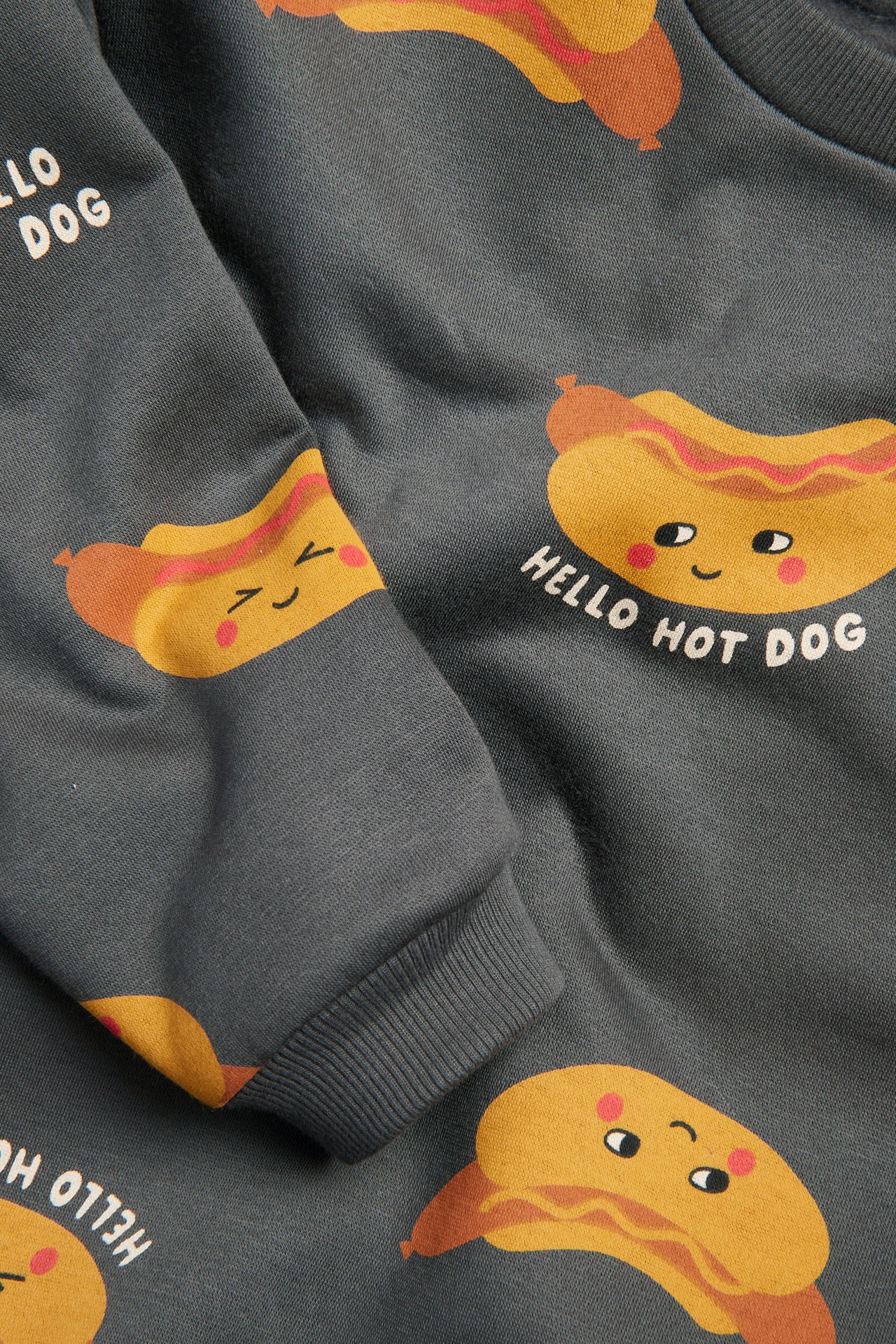 Nærbilde av en mørkegrå bomullsgenser med muntre "Hello Hot Dog"-trykk og ribbestrikkede mansjetter.