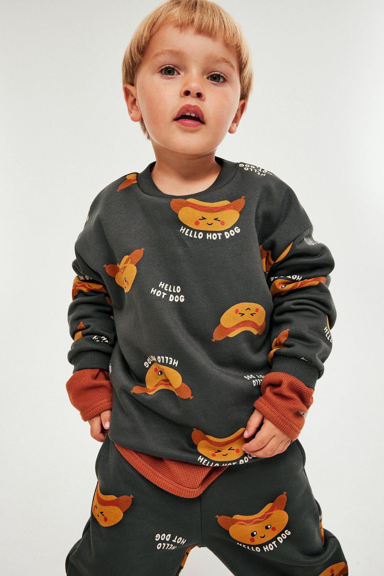 Frontalansikt: Barn i mørkegrå sweatshirt med hot dog-print, oransjebrune mansjetter og joggebukse.