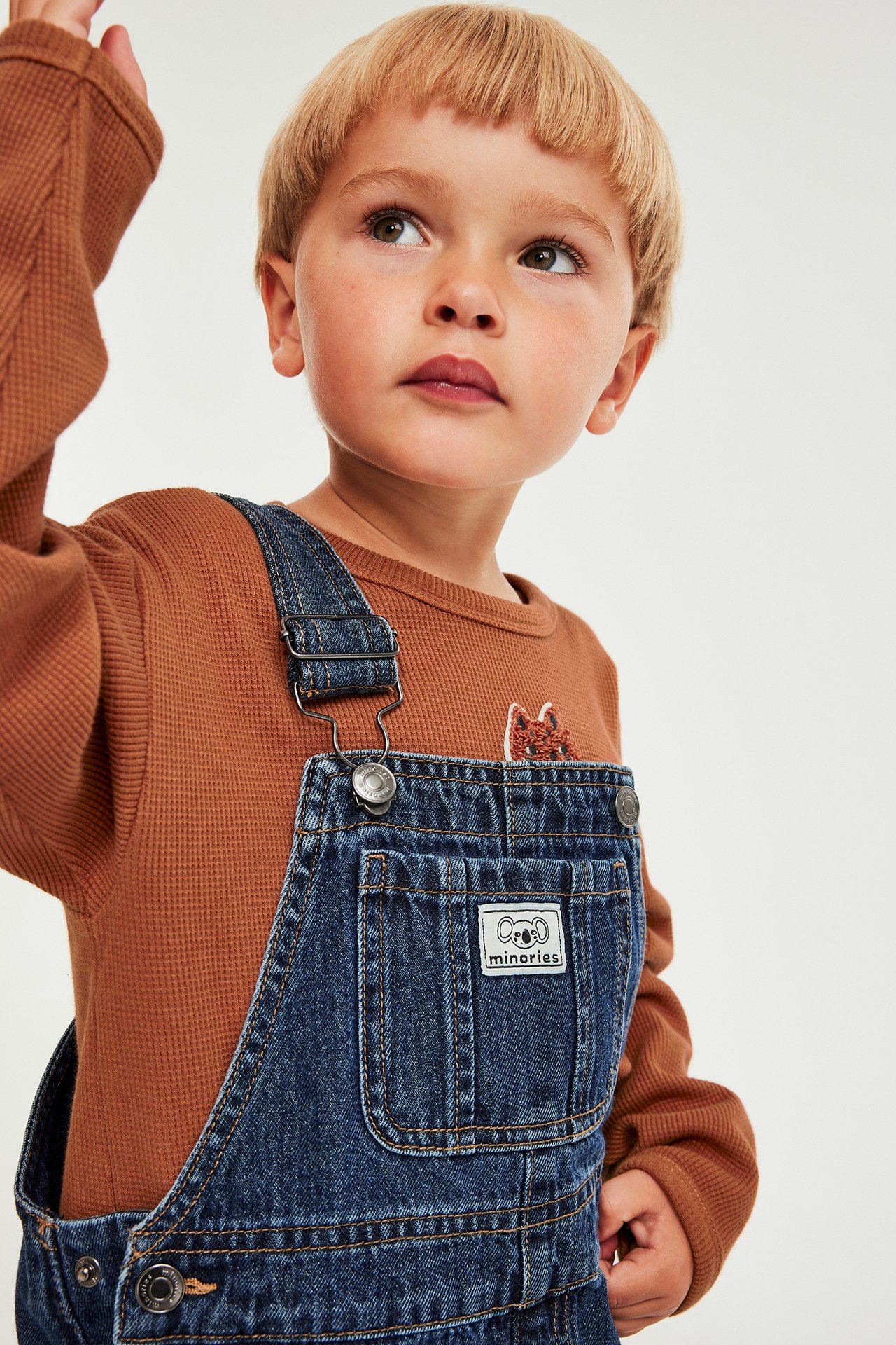 Barnpojke i brun långärmad topp med brodyr, sportig denim overall, frontvy.