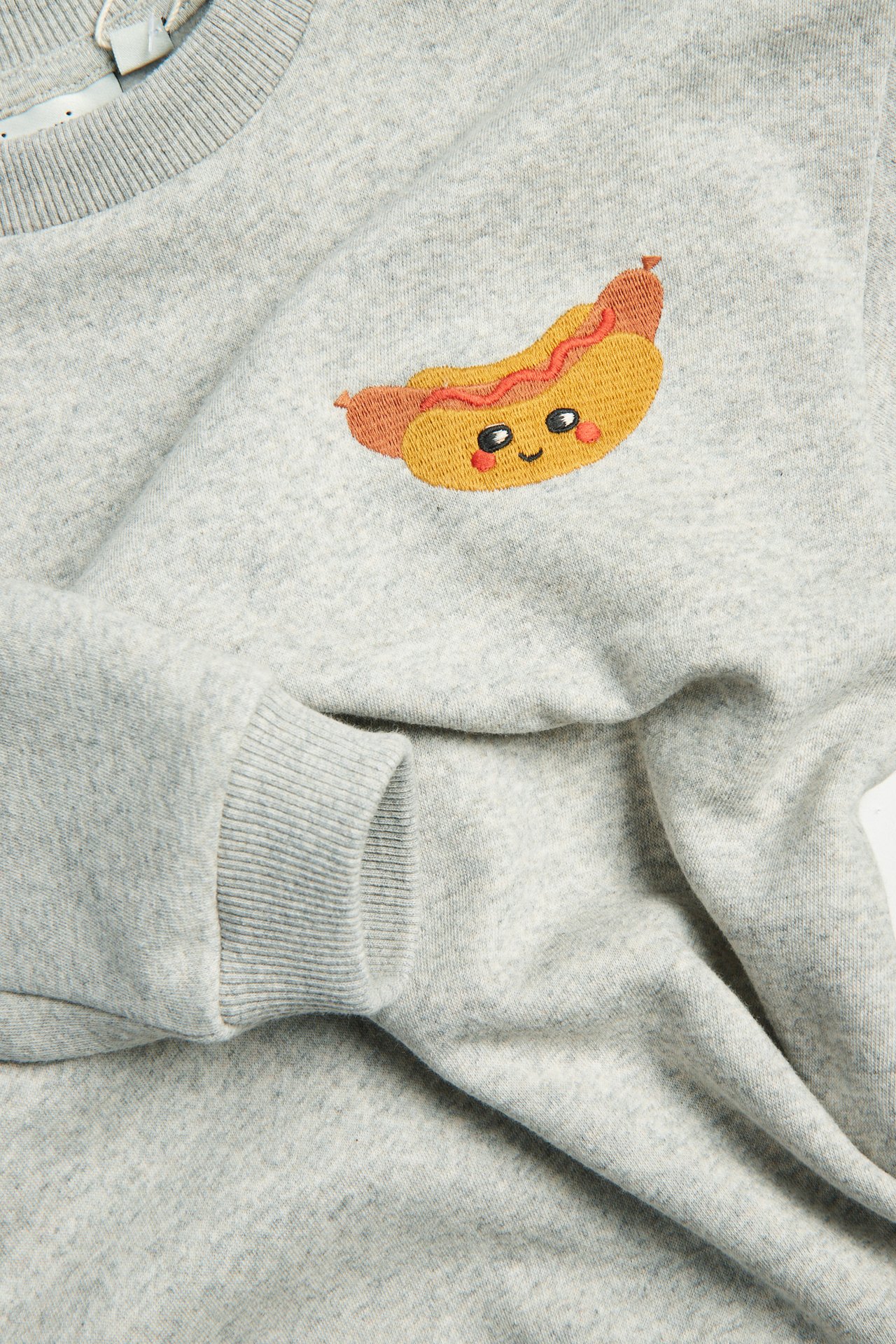 Nærbilde forfra av en lys grå barnesweatshirt med brodert pølse på brystet.