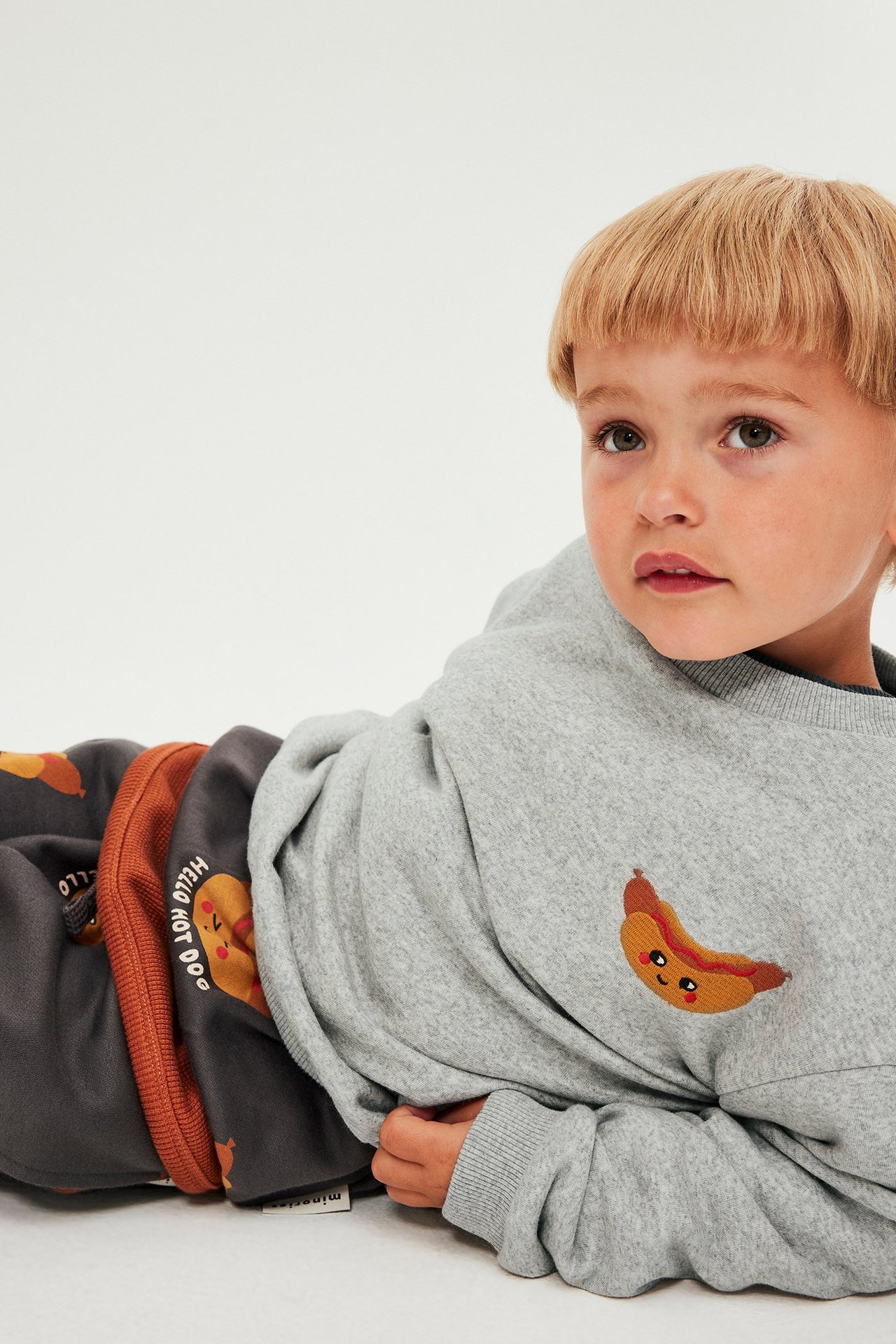 Forfra: barn iført lysegrå sweatshirt med brodert pølse, ved siden av stablede gensere.