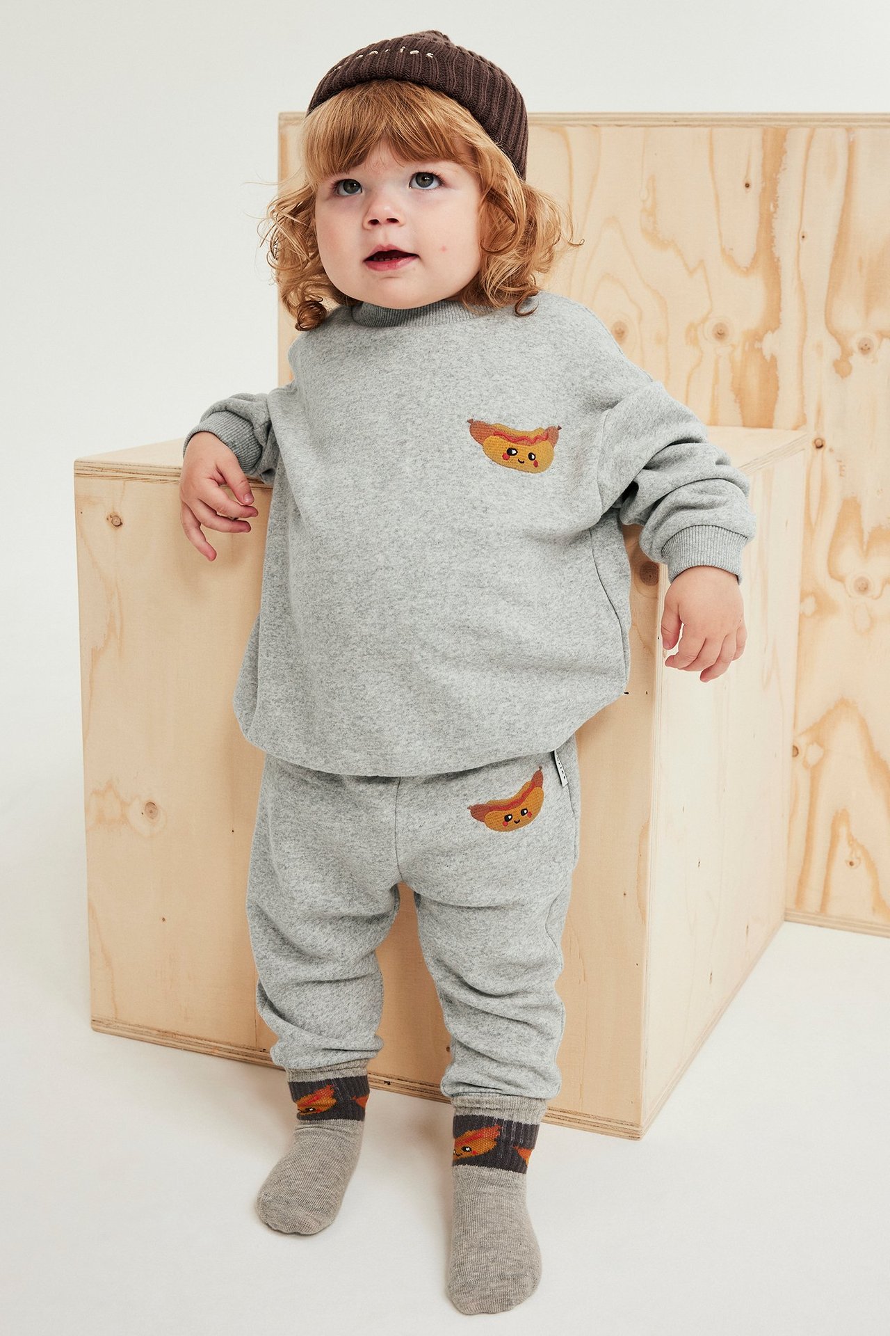 Frontalt bilde av en baby i lysegrå joggebukse med pølsebroderi, matchende genser, brun lue og mønstrede sokker.