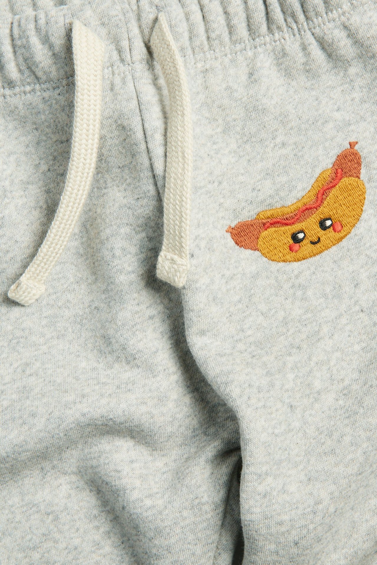 Nærbilde av lysegrå babyjoggebukse med en brodert pølse-design og hvit snøring.