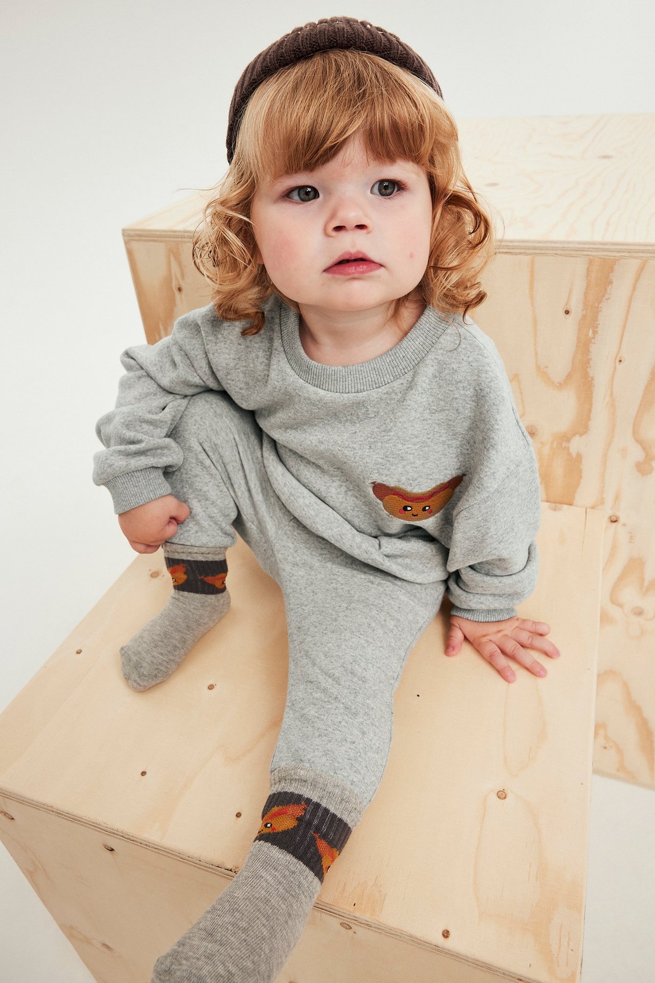 Høyvinkelbilde av baby i lys grå joggedress med pølsebroderi, brun lue og mønstrede sokker.