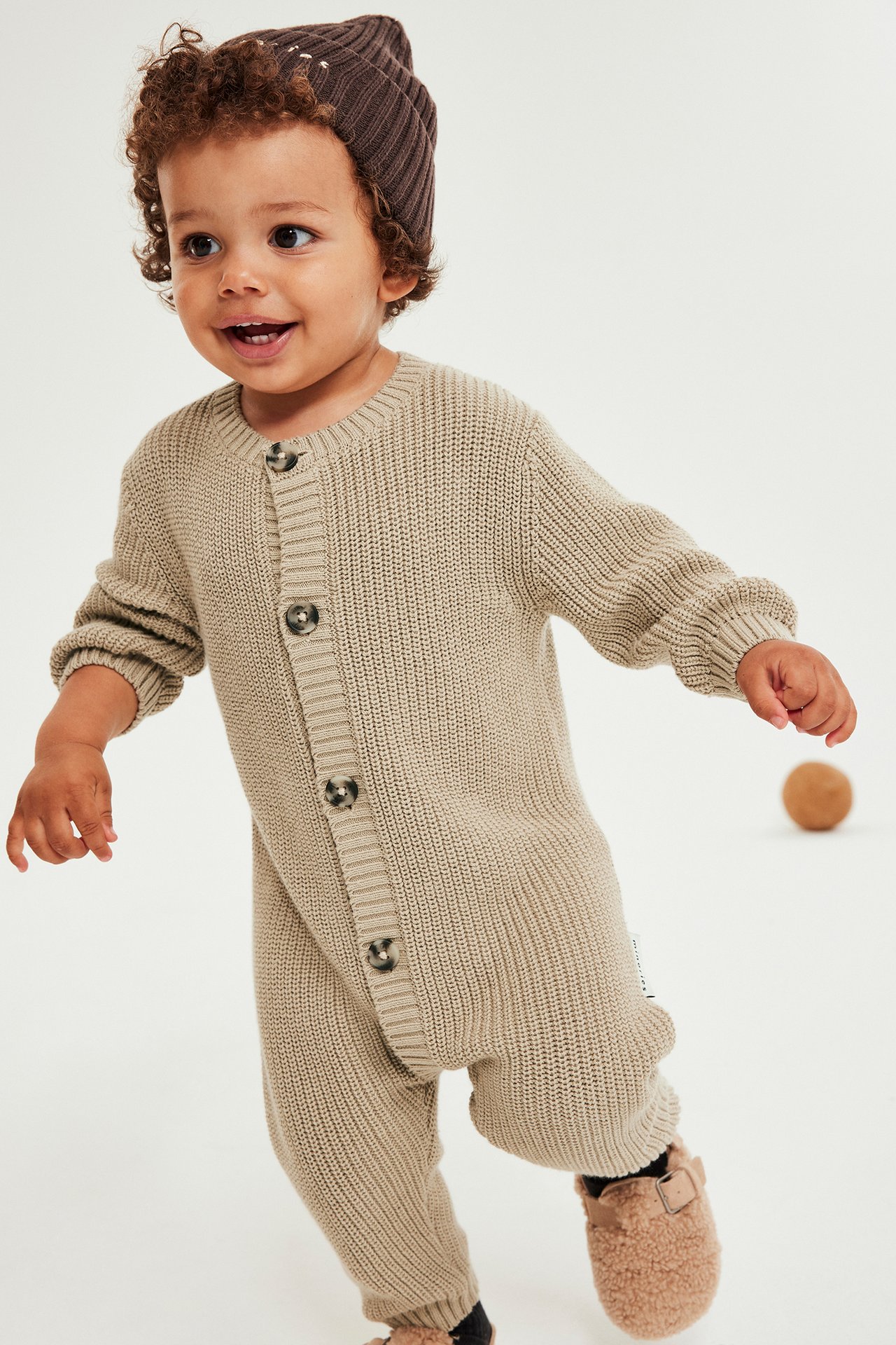 Beige stickad baby jumpsuit med knappar fram och långa ärmar, bärs av liten pojke.
