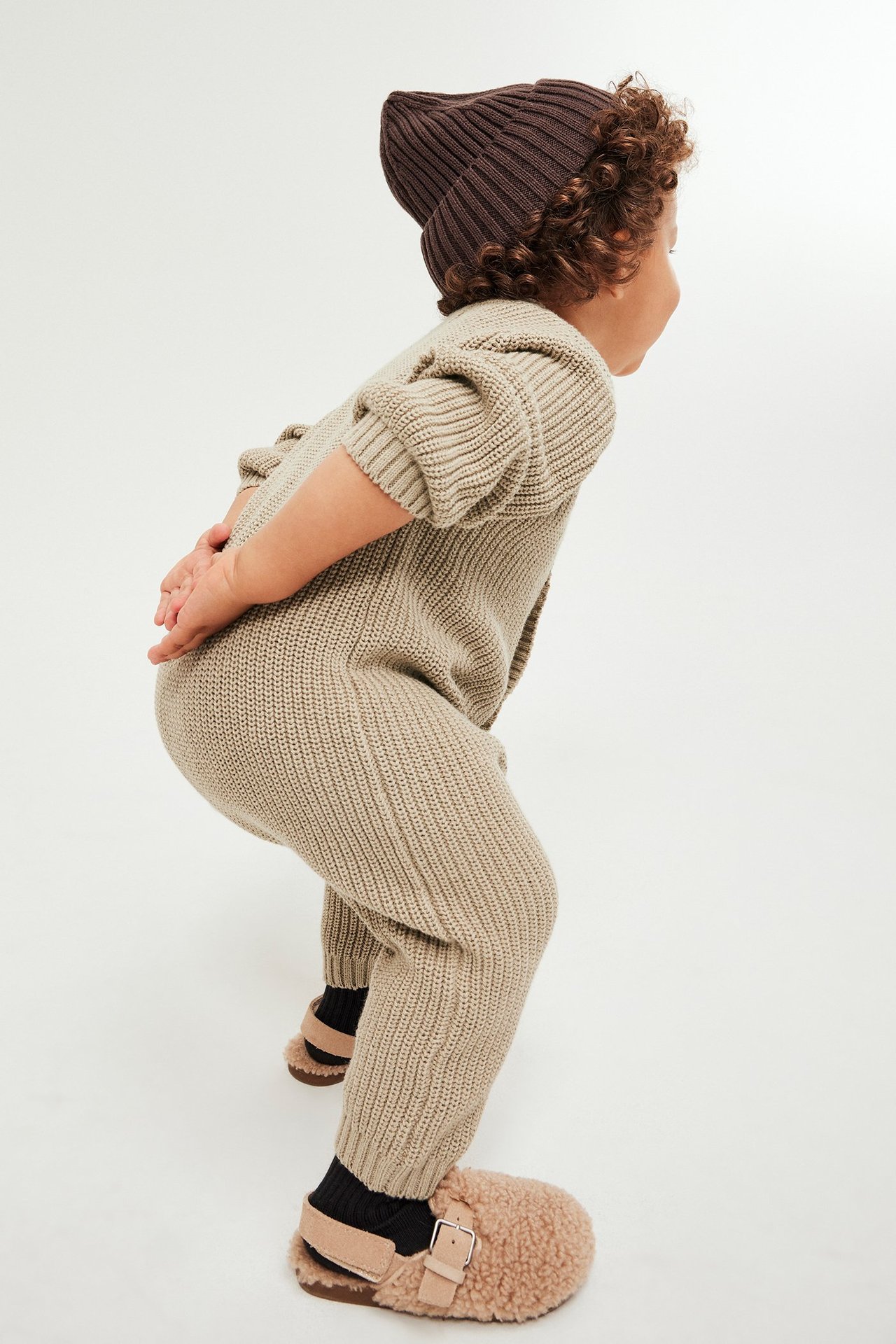 Beige stickad baby jumpsuit med knappar framtill, sett från sidan, på liten pojke.