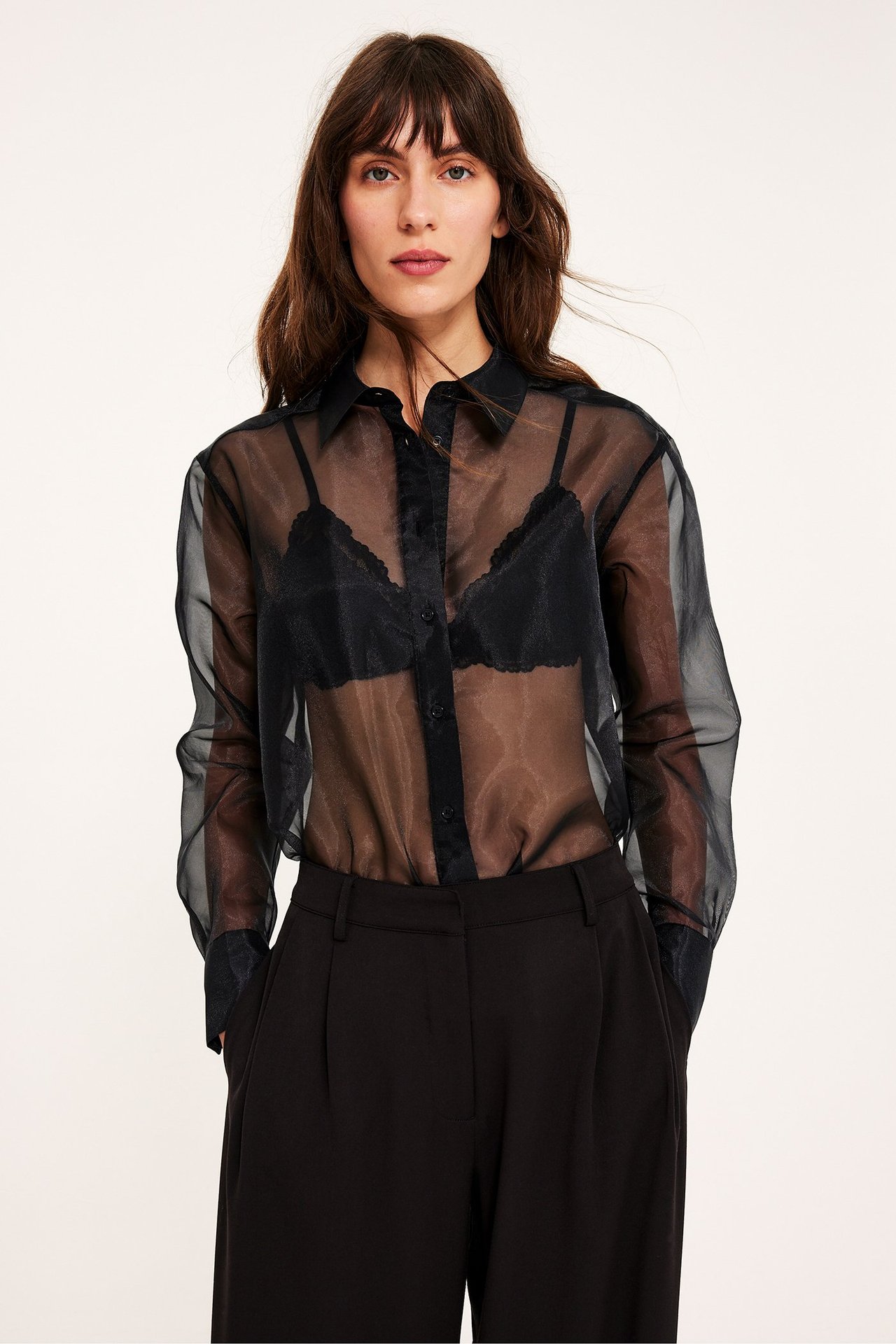 Framifrån, kvinna i svart transparent organzaskjorta över svart BH och svarta byxor.