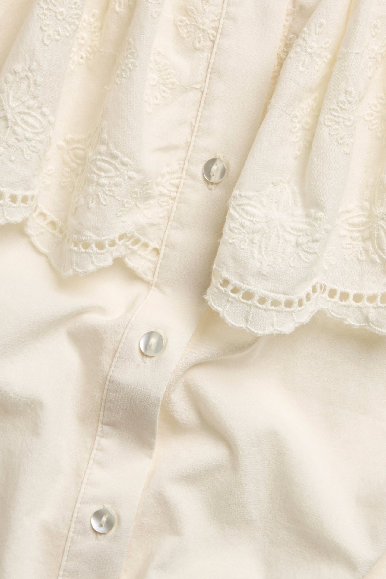 Zbliżenie na ecru bawełnianą bluzkę z haftowaną peleryną w kwiaty i guzikami z przodu.