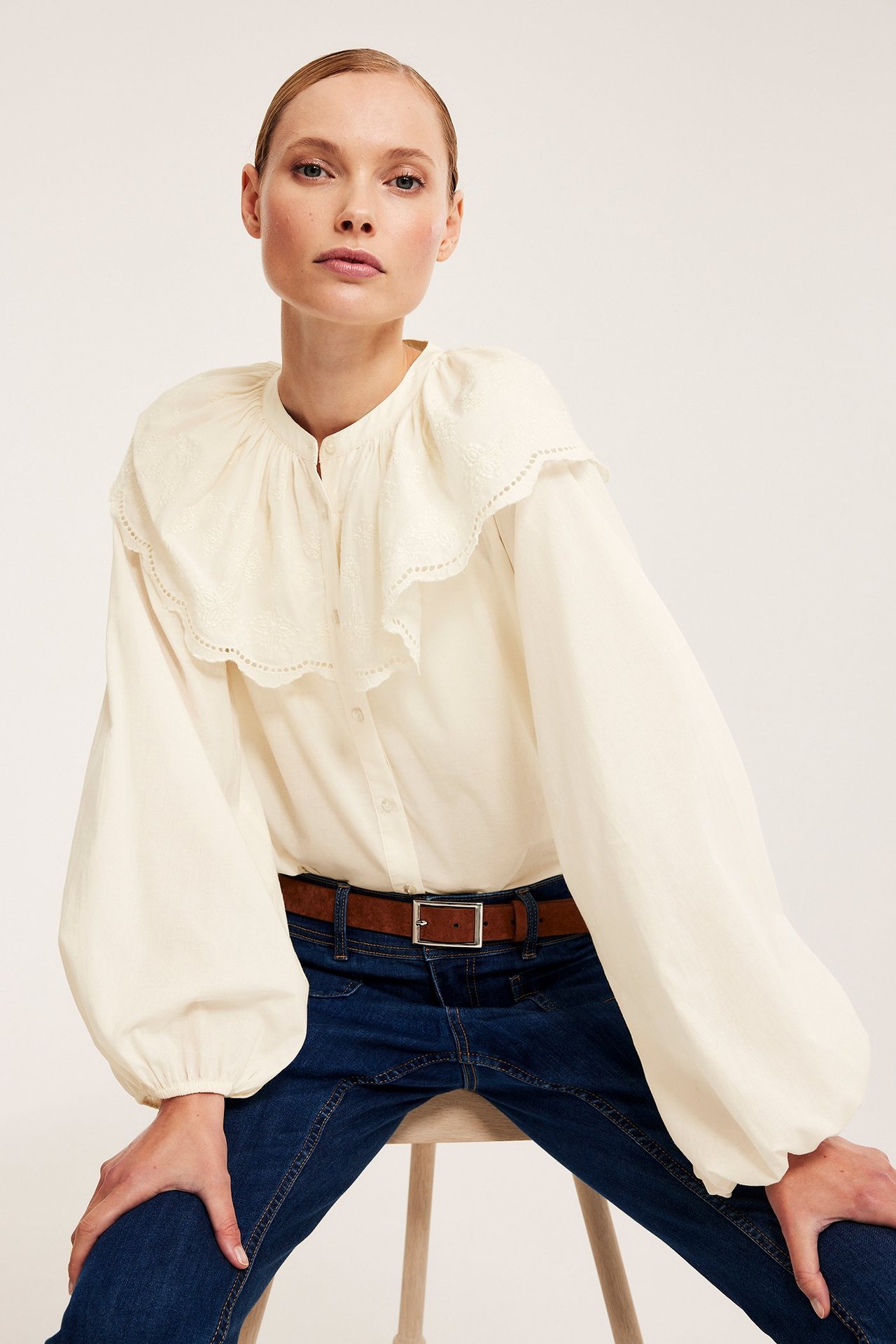 Edestä, nainen luonnonvalkoisessa puserossa brodeeratulla capella, tummansiniset farkut ja ruskea vyö.