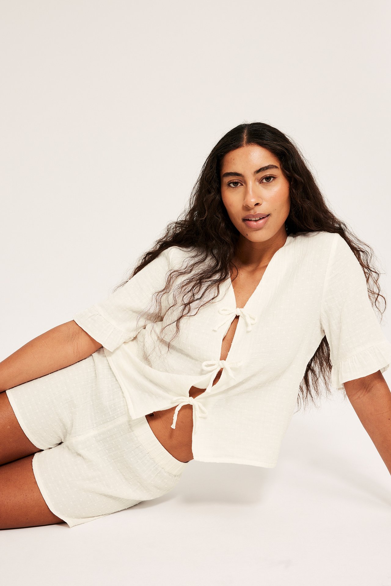 Forfra: kvinne i off-white teksturert bomullspyjamasett med knytetopp og shorts.