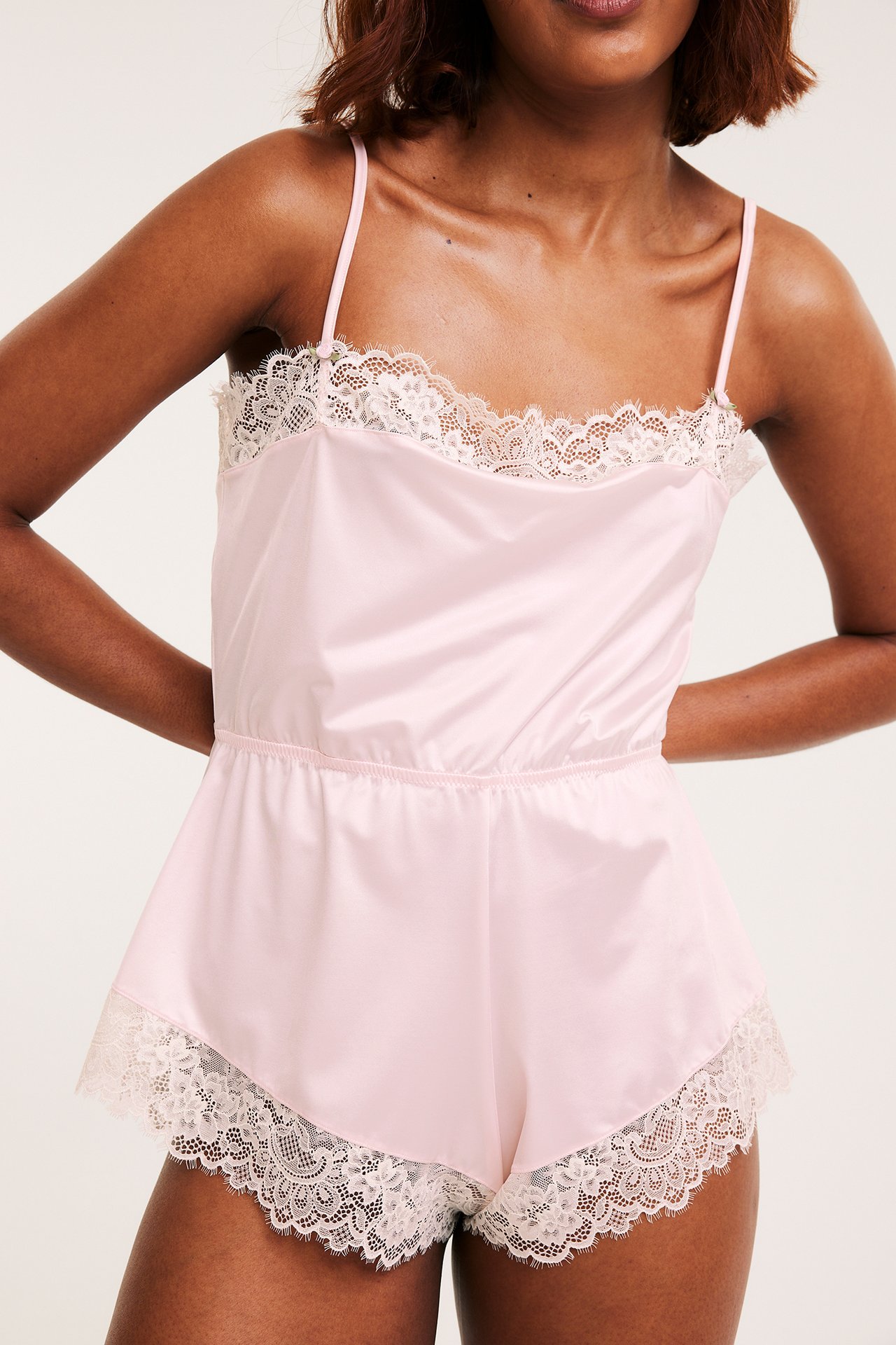 Edestä, nainen vaaleanpunaisessa satiini-playsuitissa valkoisella pitsillä ja ohuilla olkaimilla.