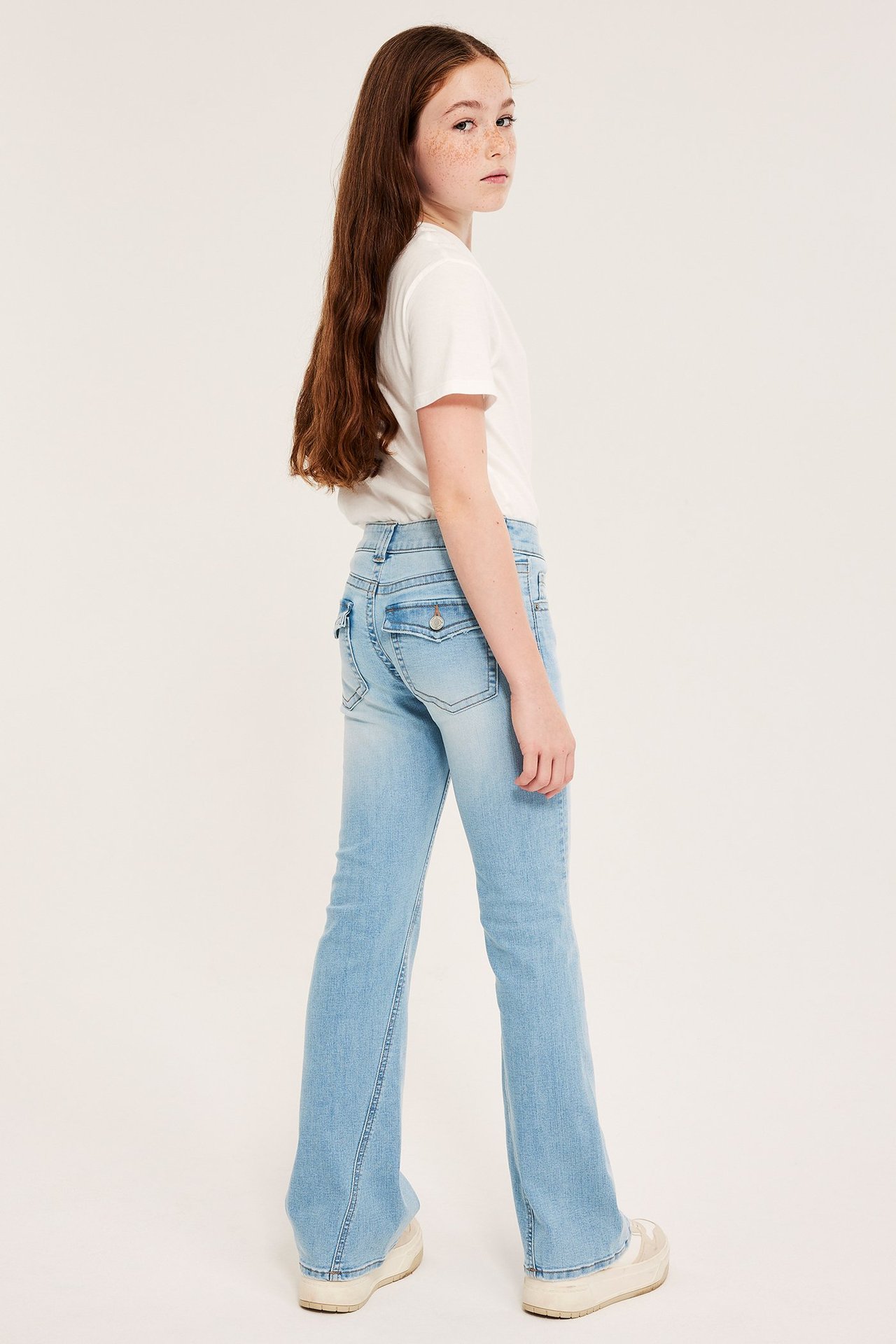 Sidestilt bilde av barn i lyseblå lavmidje bootcut-jeans, hvit t-skjorte og joggesko.