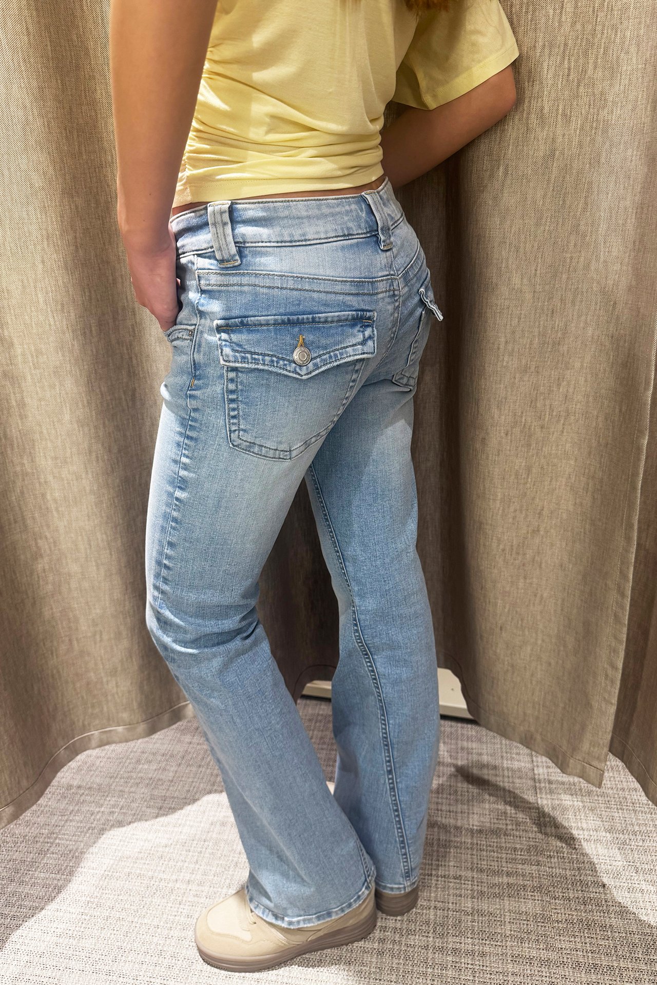 Widok z boku kobiety w jasnoniebieskich jeansach bootcut z niskim stanem, skręconymi szwami, i żółtym topie.