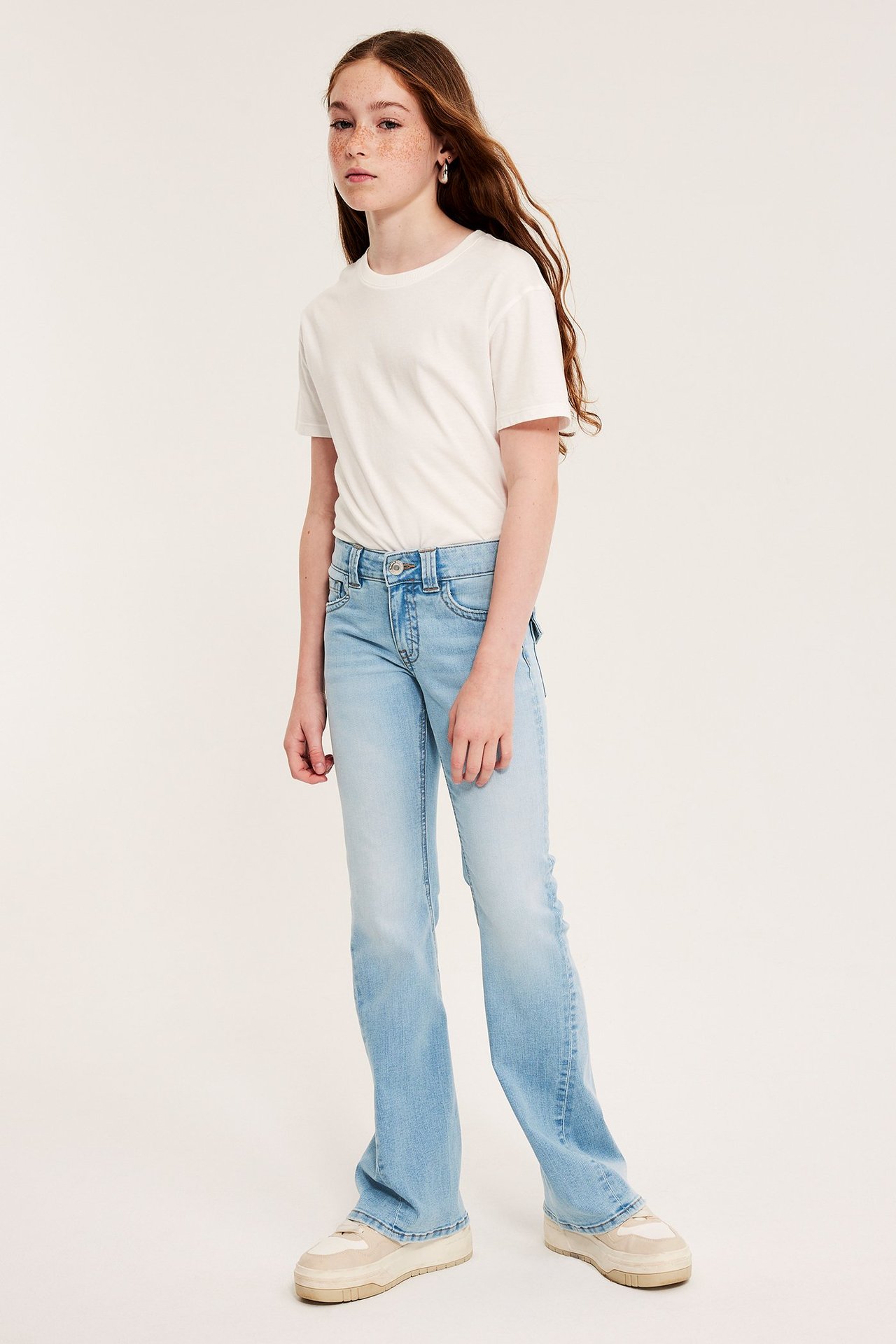 Edestäpäin: lapsi vaaleansinisissä bootcut-farkuissa, valkoisessa t-paidassa ja beigessä tennareissa.