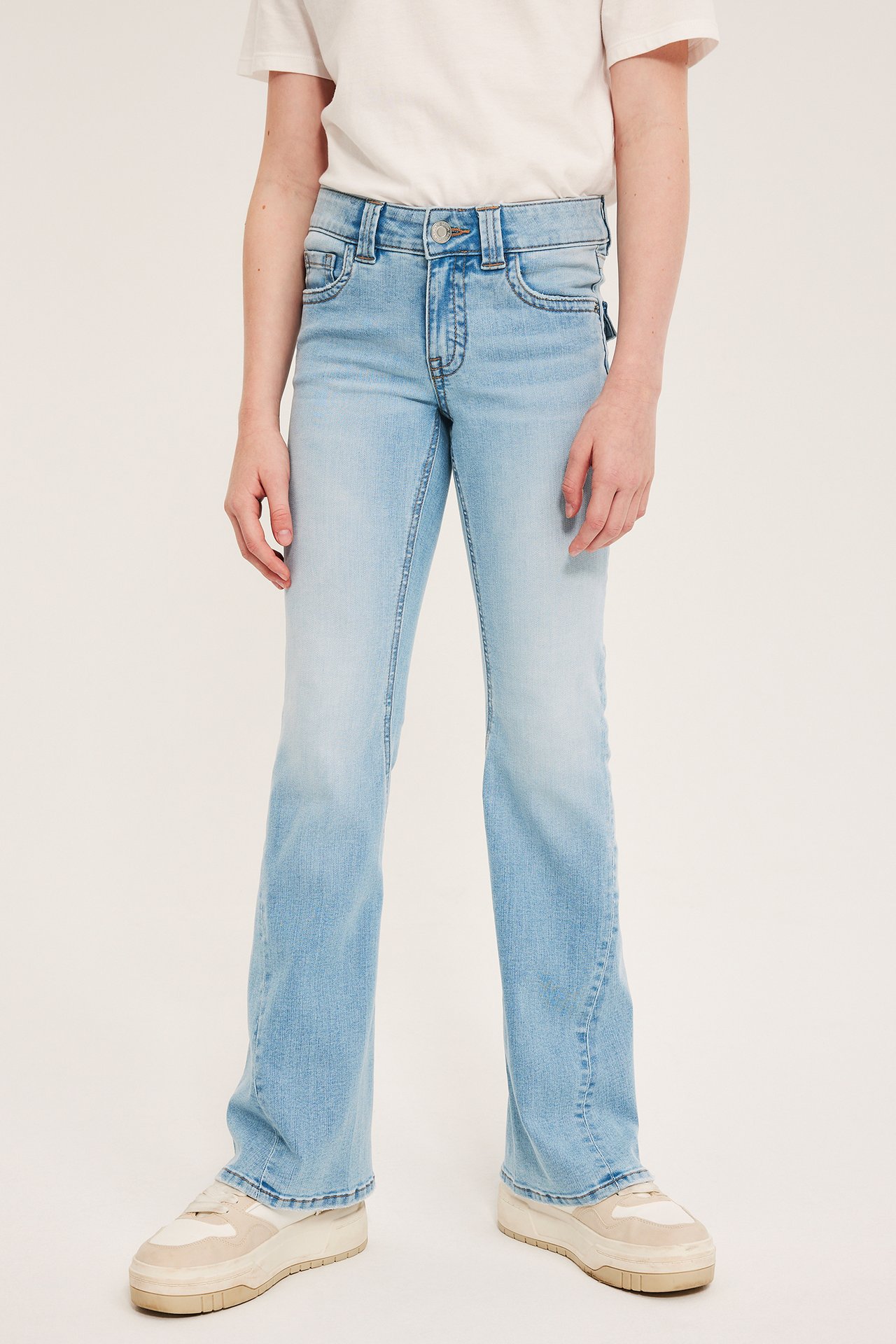 Frontbilde av et barn i lys denim bootcut-jeans med lav midje, med hvit t-skjorte og beige joggesko.