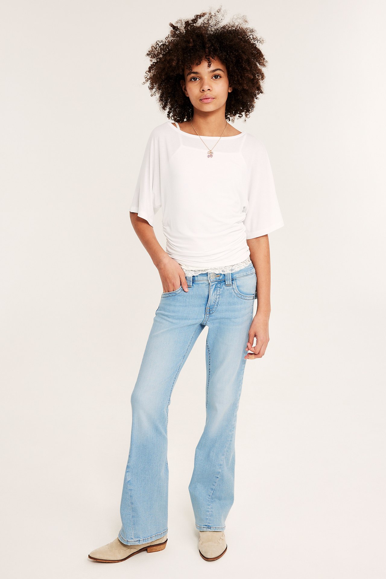 Widok z przodu: Dziecko w jasnoniebieskich jeansach bootcut z niskim stanem, białym topie i jasnobeżowych botkach.