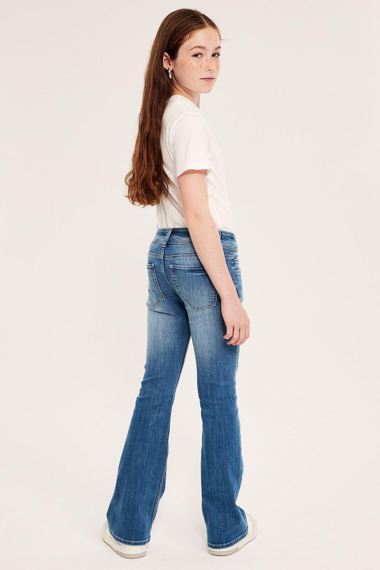 Dziecko nosi niebieskie dżinsy bootcut z niskim stanem, kopertowym pasem i biały t-shirt, widok z tyłu.