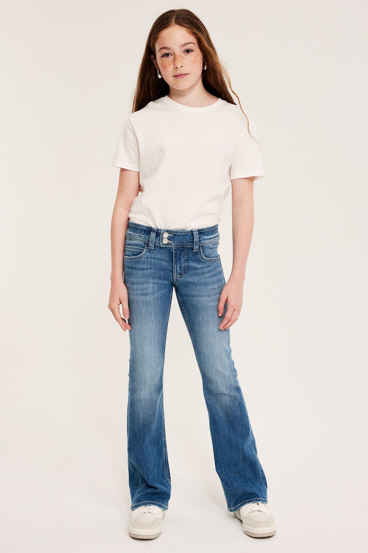 Z przodu: Dziecko w niebieskich jeansach bootcut z niskim stanem, z podwójnymi guzikami, białym T-shirtem i sneakersami.