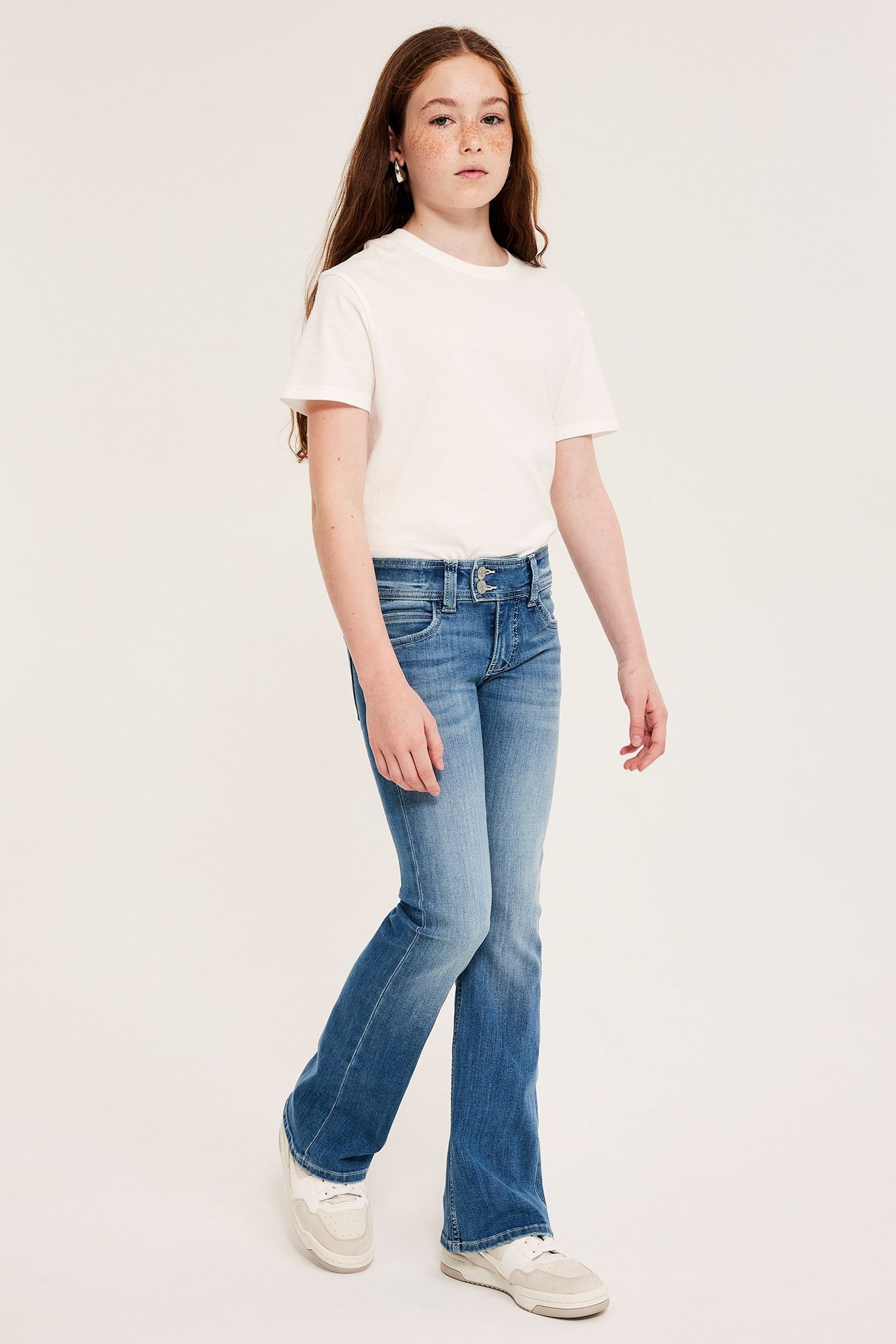 Widok z przodu: dziecko w niebieskich jeansach bootcut z podwójnym zapięciem, białym T-shircie i białych tenisówkach.