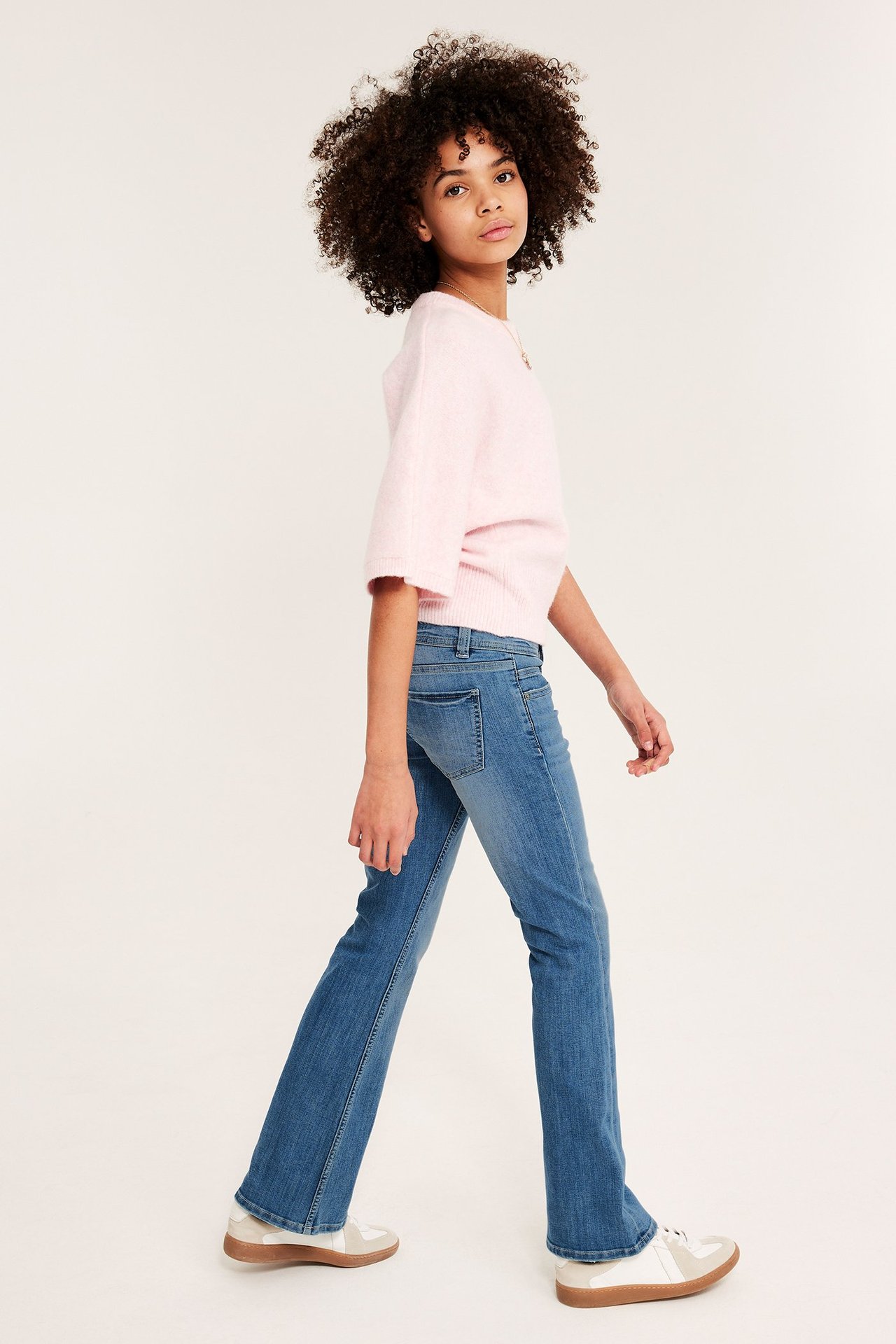 Sivukuva lapsesta, jolla on siniset matalavyötäröiset bootcut-farkut, vaaleanpunainen neule ja valkoiset tennarit.