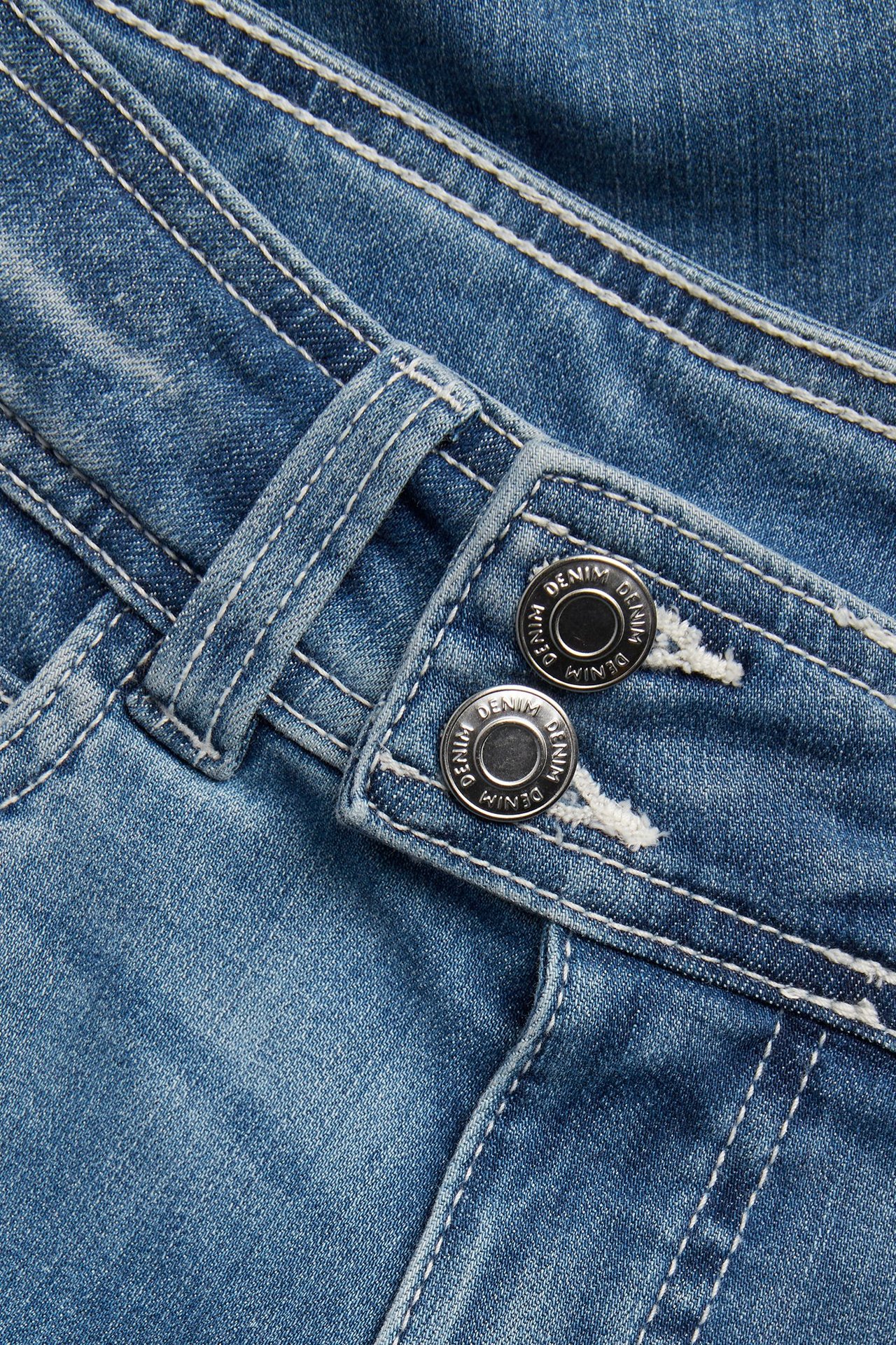 Zbliżenie niebieskich jeansów bootcut z białymi szwami i zakładanym podwójnym zapięciem na guziki w pasie.