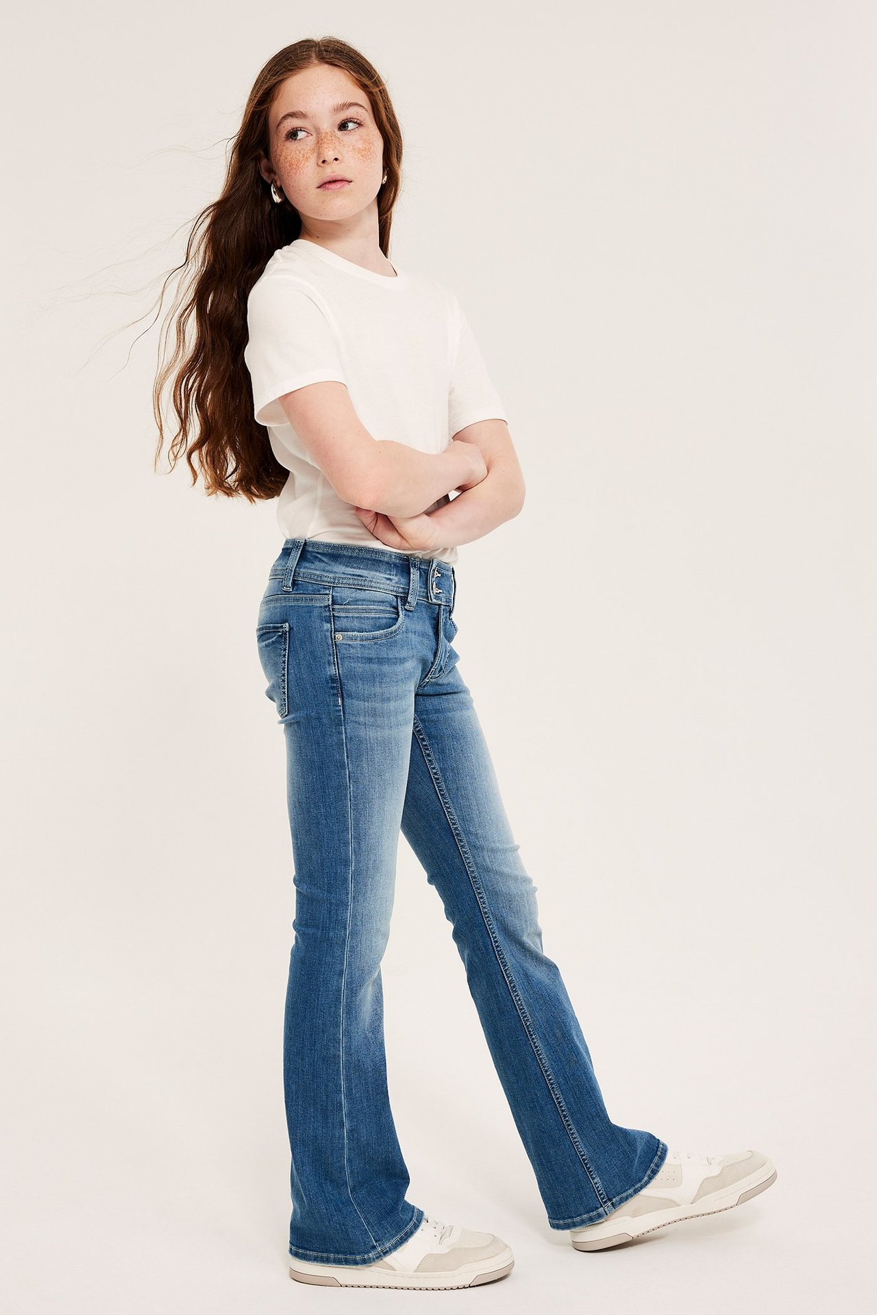 Widok z boku: dziecko w niebieskich dżinsach bootcut z podwójnym guzikiem, białym t-shircie i trampkach.