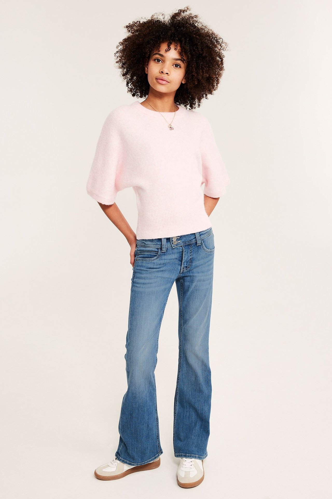 Dziewczynka w niebieskich dżinsach bootcut z zakładką, jasnoróżowym swetrze i trampkach.