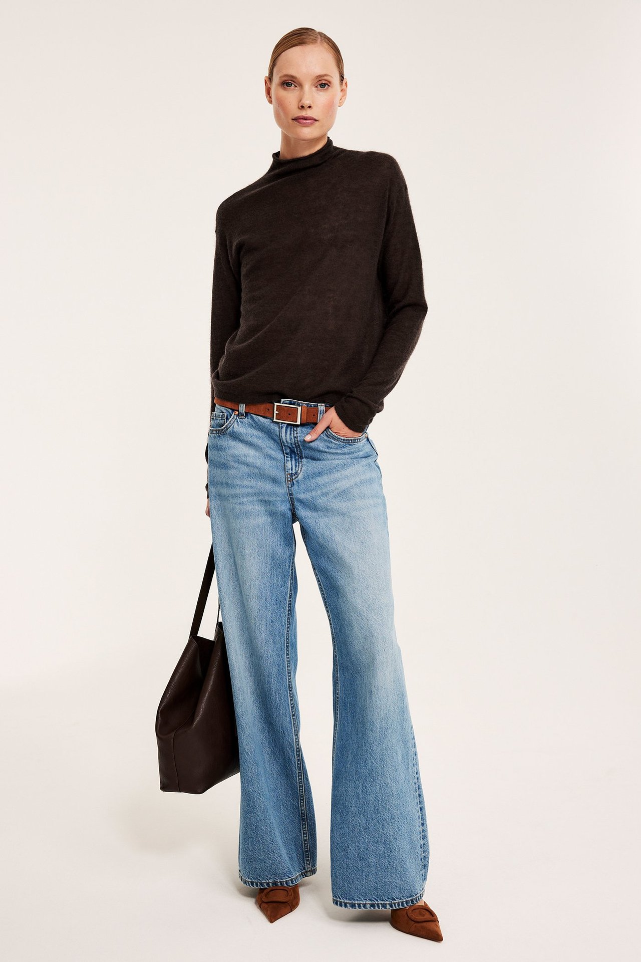Frontbilde, kvinne i mørkebrun melert ullgenser, lyseblå jeans, brunt belte og veske.