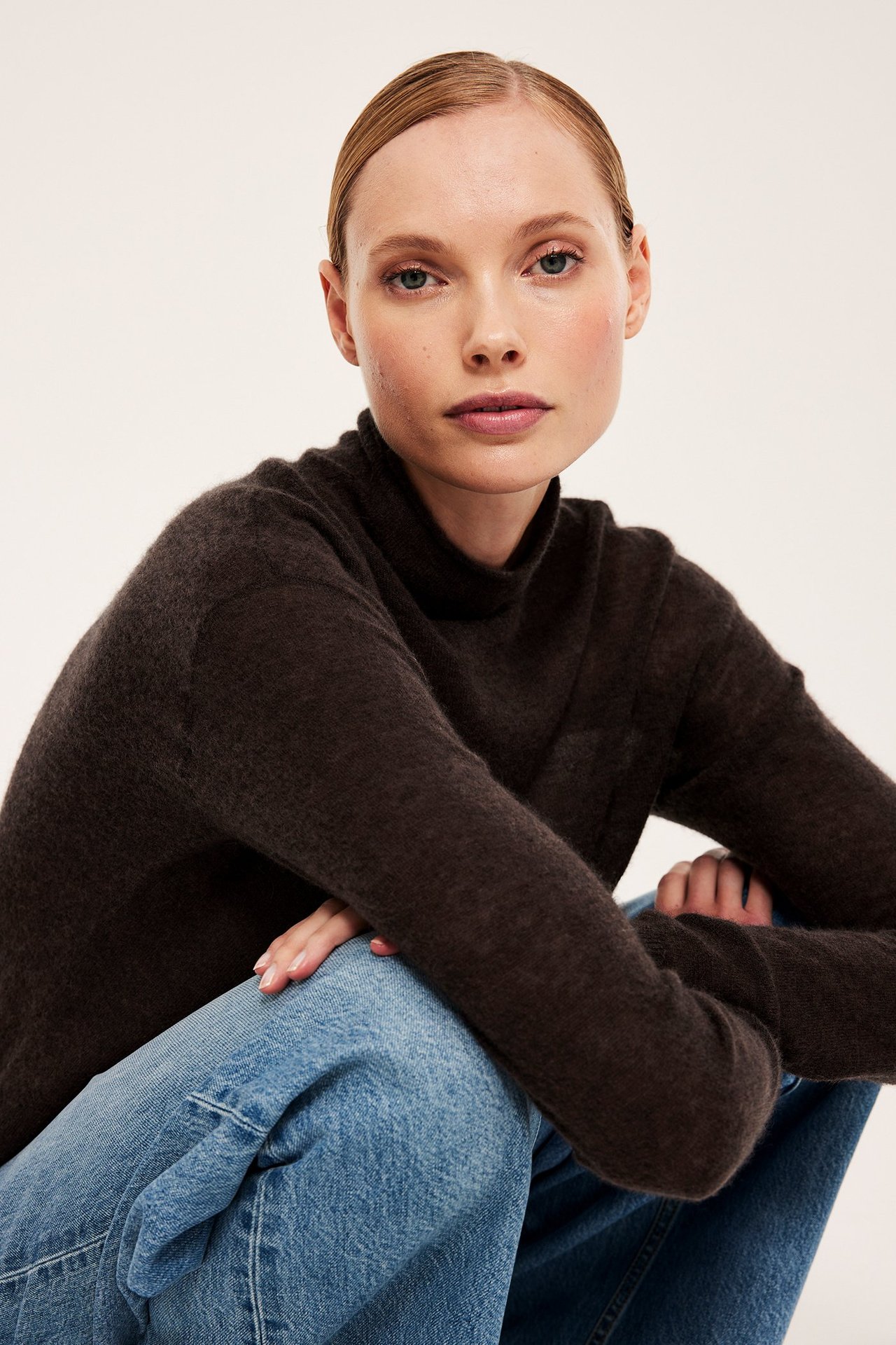 Forfra: kvinne i en tynn, mørkebrun ullblanding turtleneck-genser og lyseblå jeans.