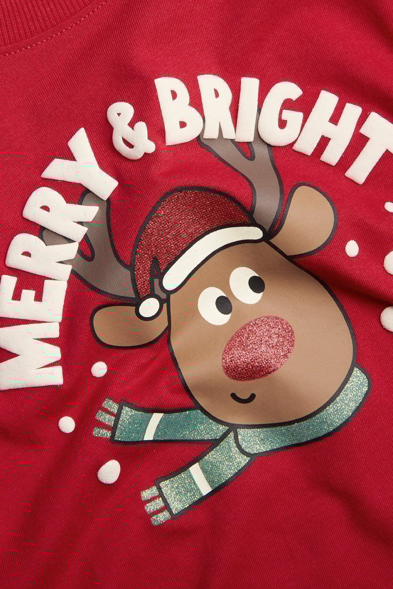 Zbliżenie czerwonej dziecięcej bawełnianej koszulki z długim rękawem, z nadrukiem Rudolfa i 'Merry & Bright'.