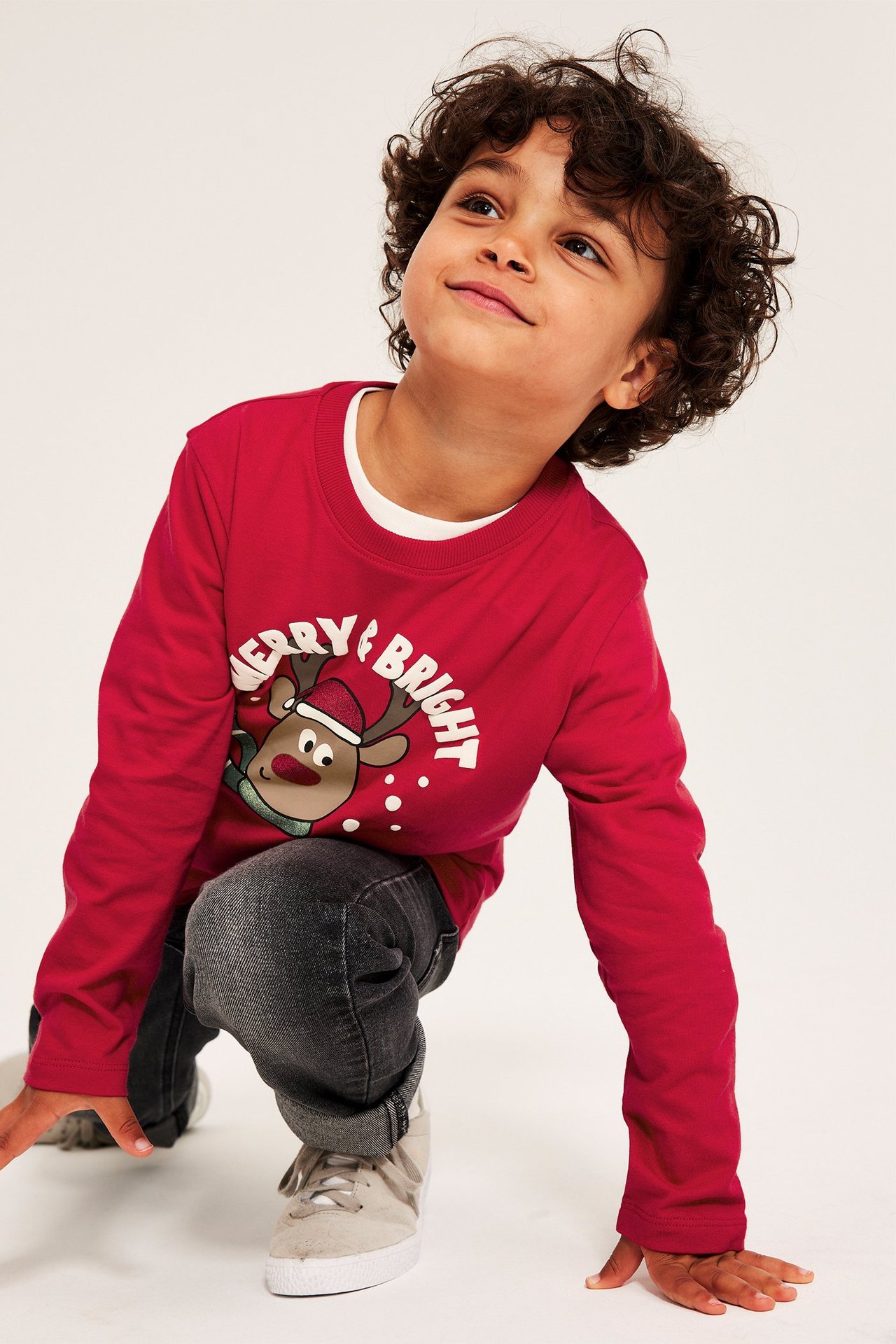 Framifrån: Barn i röd långärmad t-shirt med Rudolf-tryck, grå jeans och beiga sneakers.
