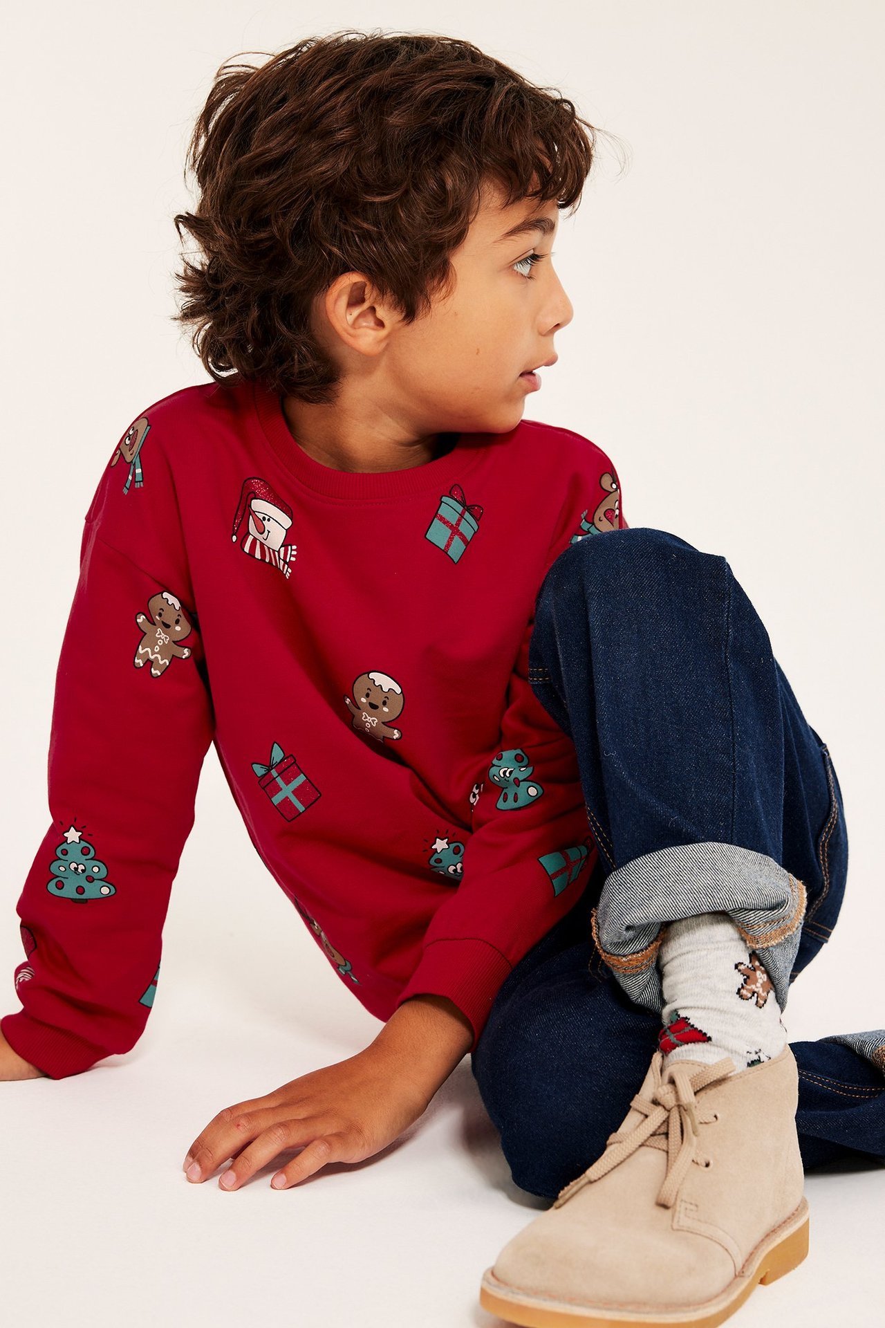 Barnkille i rödfärgad sweatshirt med julmotiv, blå jeans och beige boots, sedd i sidovyn.
