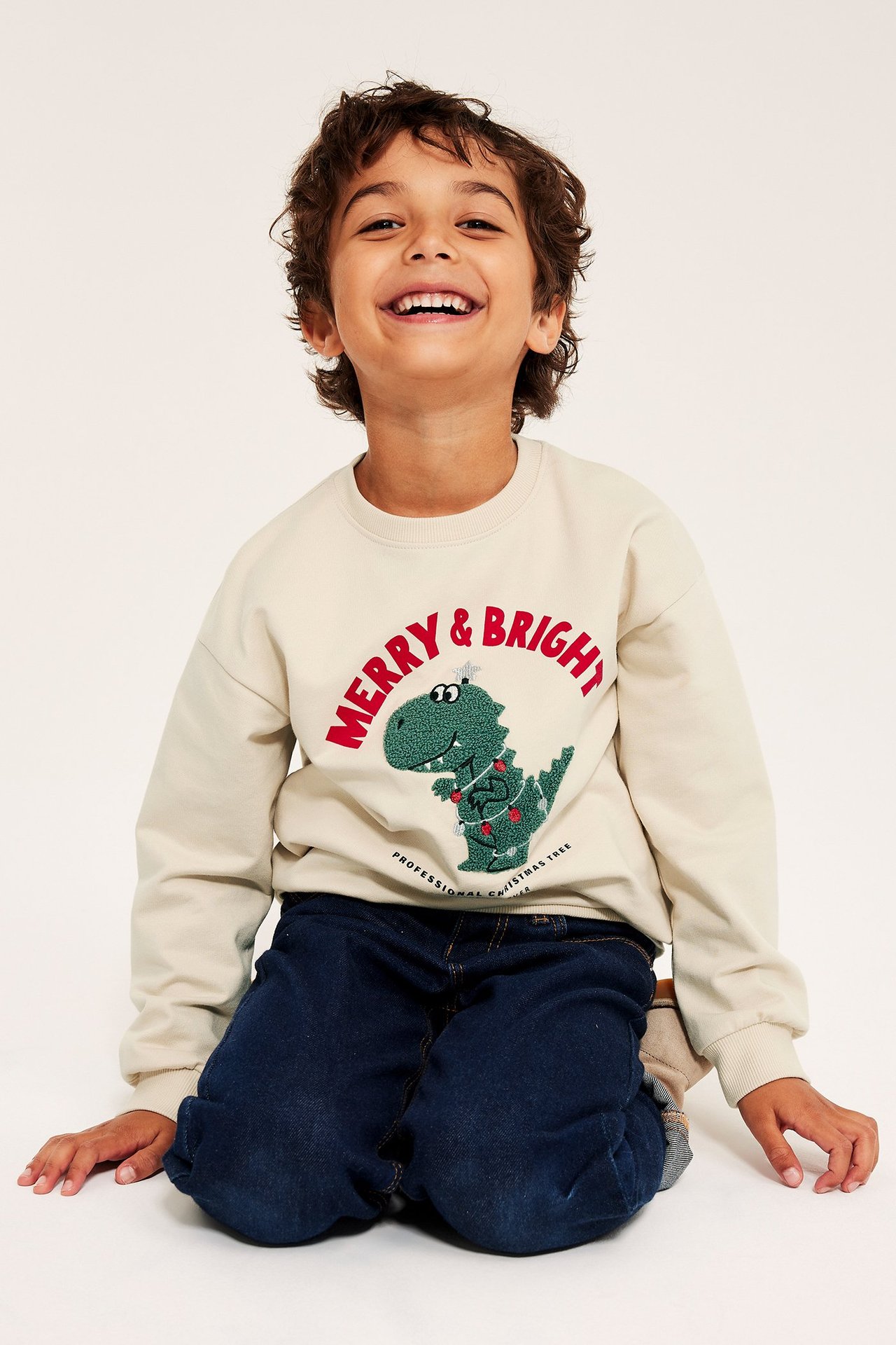 Barns sweatshirt i ljusbeige med dinosauriemotiv och julkänsla, tryck fram, ribb i hals, ärm och nederkant.