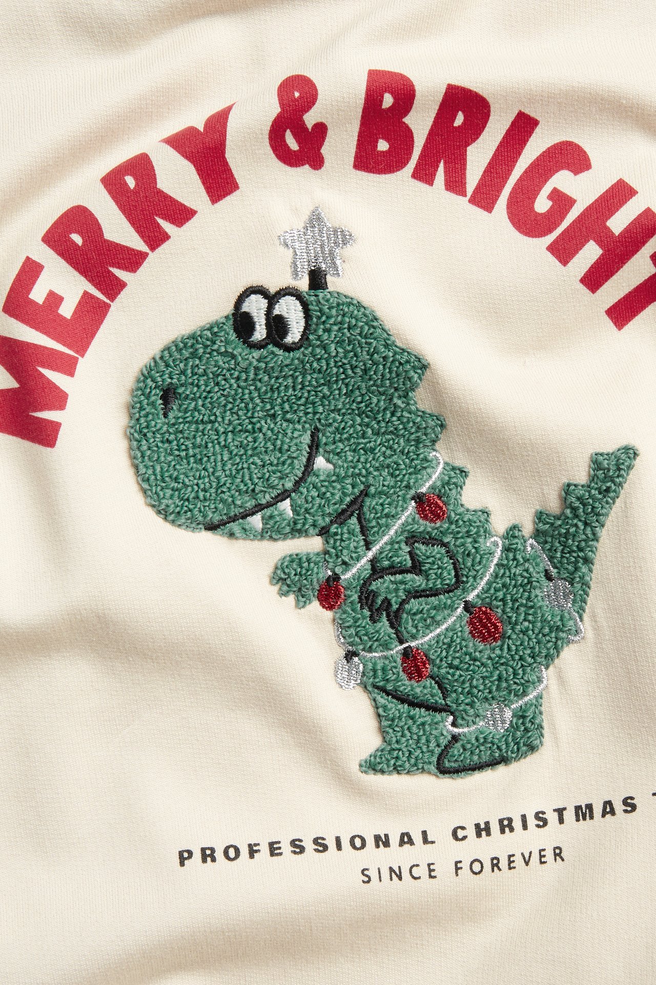 Nærbilde: Lys beige bomullsgenser for barn med grønt juledinosaur-trykk og "MERRY & BRIGHT"-tekst.