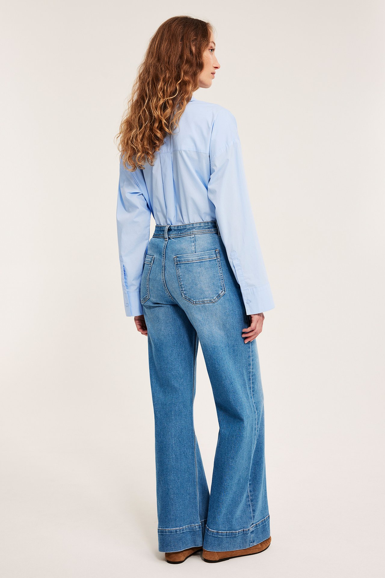 Widok z tyłu/boku kobiety w jasnoniebieskich jeansach high-waist z szerokimi nogawkami i jasnoniebieskiej koszuli.