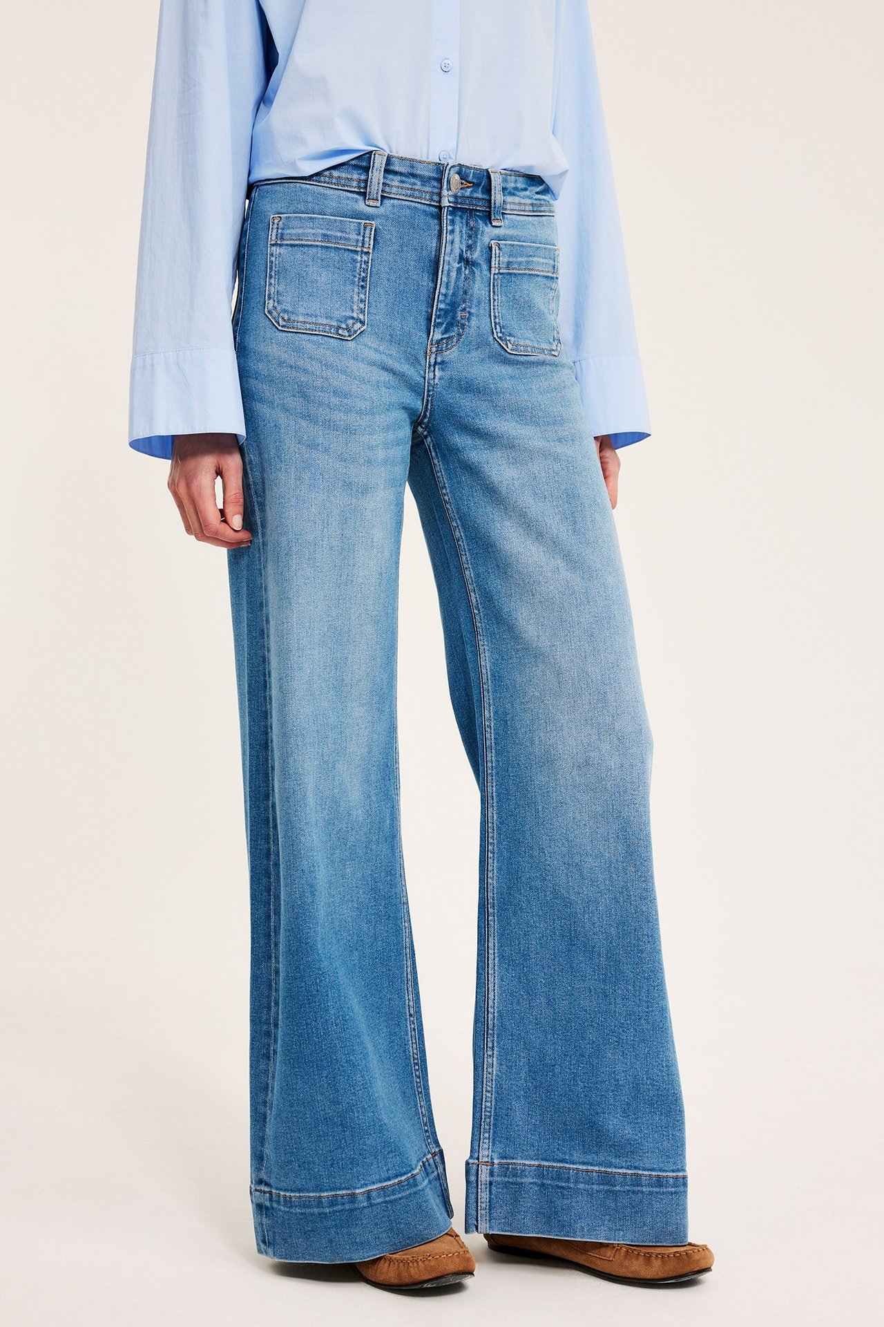 Forfra, kvinne i lyseblå vide high-waist jeans, lyseblå skjorte og brune sko.