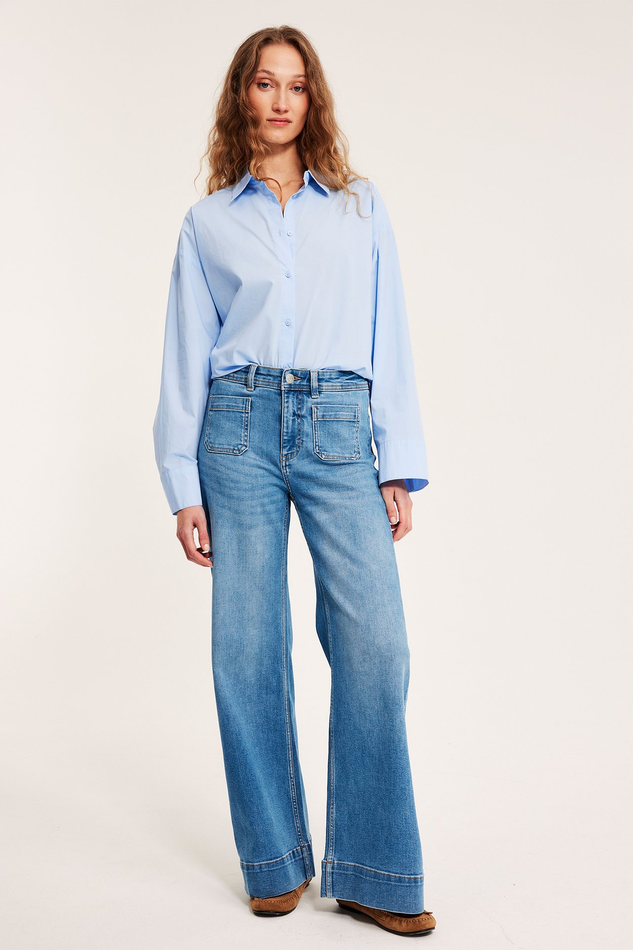 Kobieta w jasnoniebieskich szerokich jeansach z wysokim stanem i naszywanymi kieszeniami, jasnoniebieskiej koszuli i brązowych mokasynach.