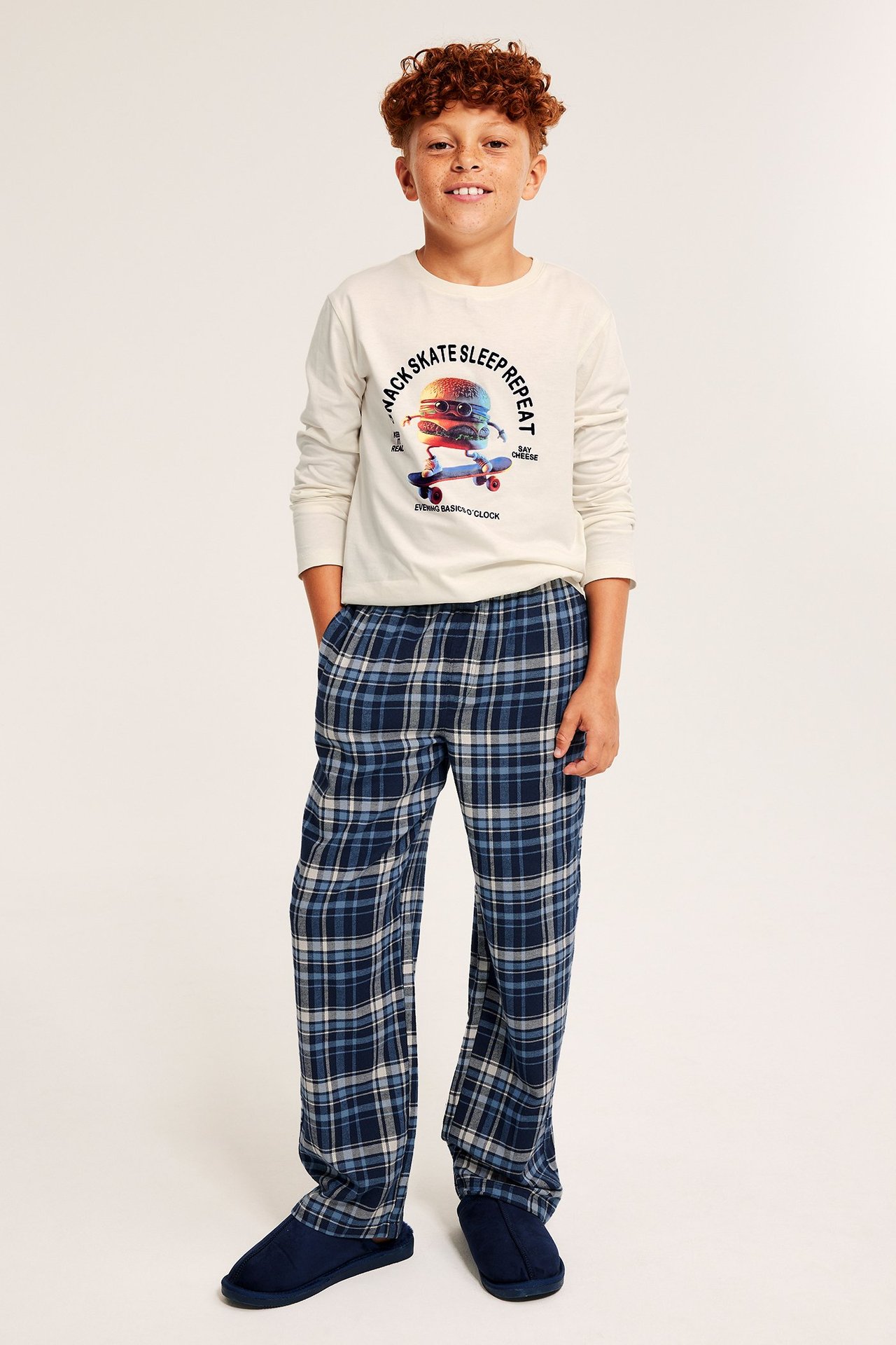 Framifrån: Barn i off-white pyjamas med tryckt topp, blårutiga flanellbyxor och blå tofflor.