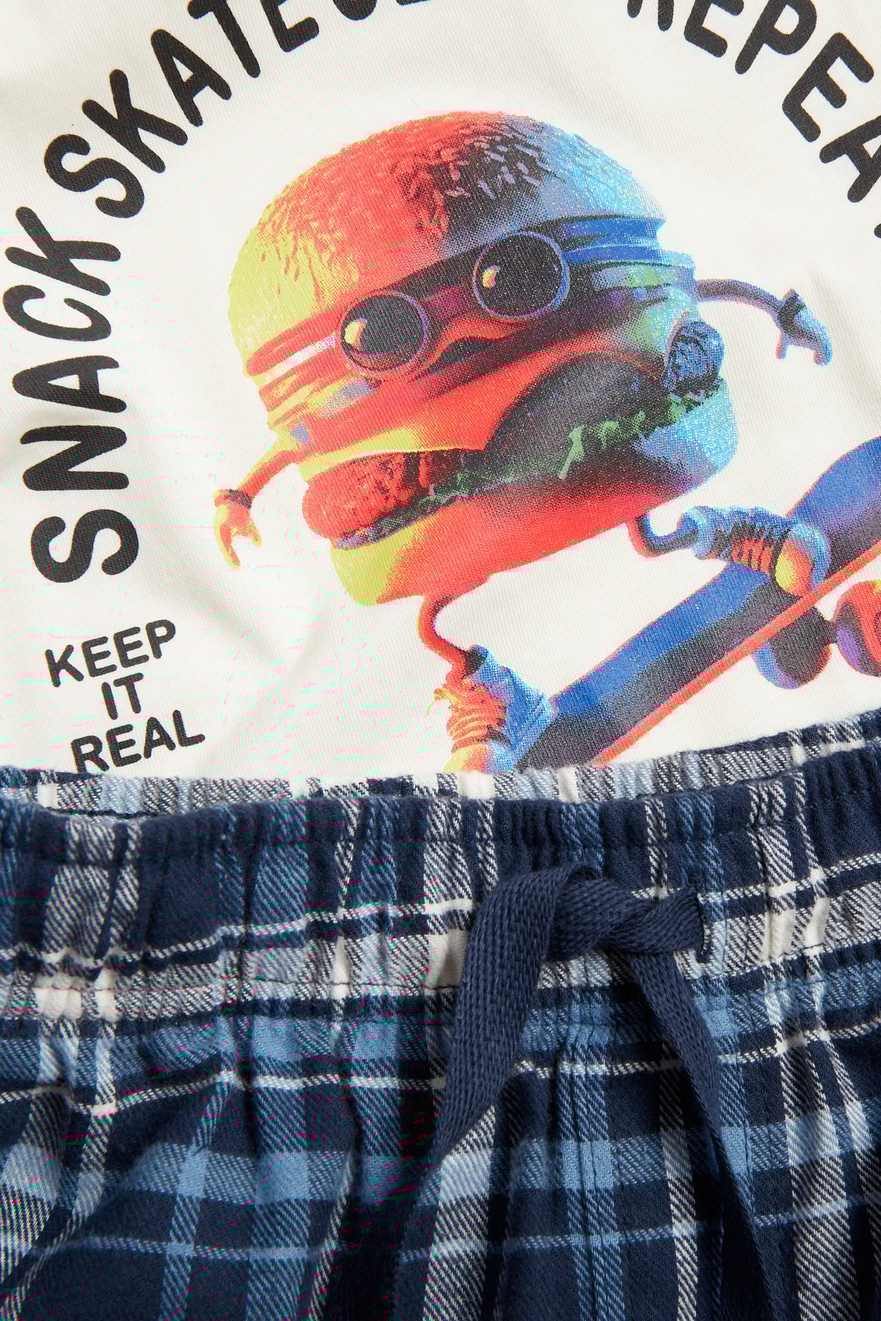 Närbild av barns offwhite pyjamaströja med burger-tryck, ihopparad med blårutiga flanellpyjamasbyxor.