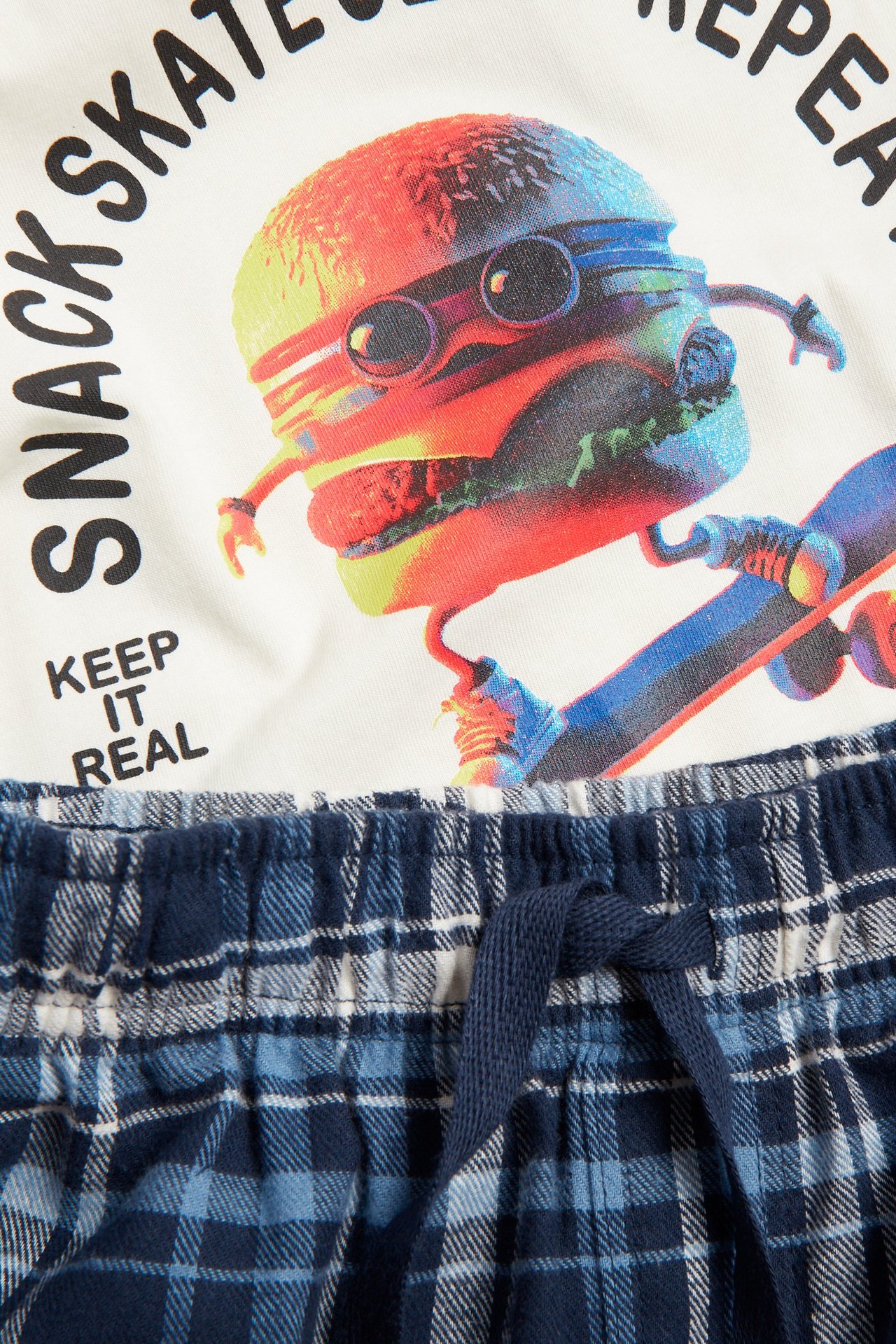 Zbliżenie na dziecięcą piżamową bluzkę w kolorze złamanej bieli z grafiką burgera, połączoną z niebieskimi, kraciastymi spodniami.