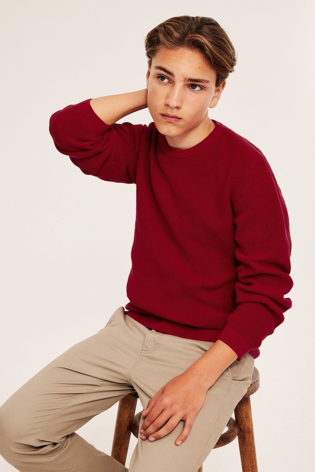 Widok z przodu: Chłopiec w ciemnoczerwonym, waflowym swetrze bawełnianym i jasnobeżowych spodniach, siedzący.