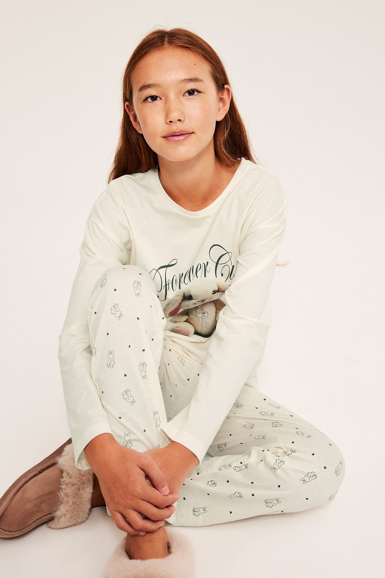 Forfra: barn i benvit bomullspyjamas; langermet topp med kaninmønster, mønstrede bukser, pelskledde tøfler.