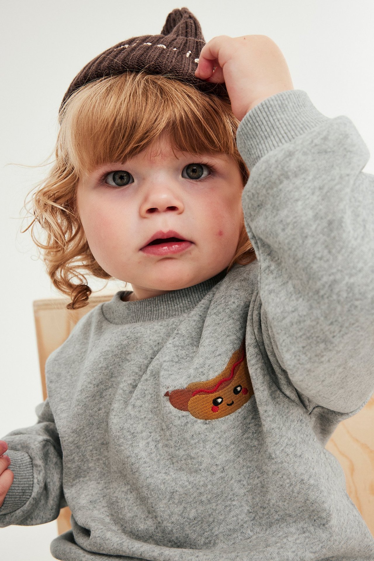 Närbild på baby i ljusgrå sweatshirt med hotdog-brodyr och en brun stickad mössa.