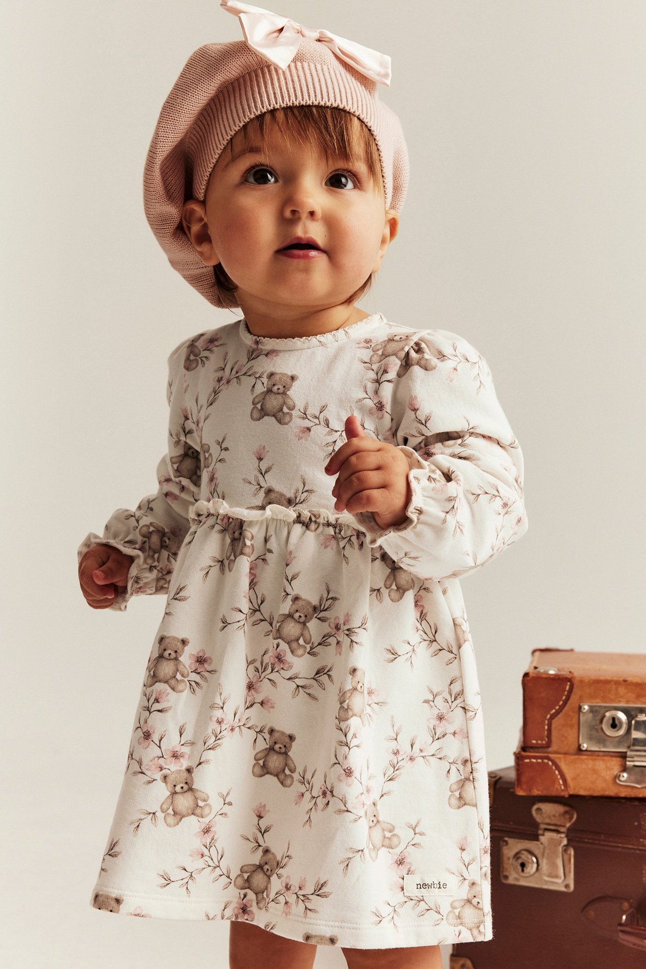 Front: Baby i kremhvit blomstret kjole med teddybjørnprint og rosa beret med sløyfe.