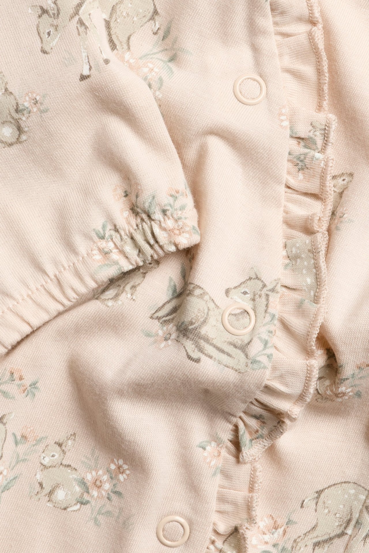 Närbild av en rosa baby-romper i bomullsjersey med rådjurs- och blommönster, volanger, tryckknappar.