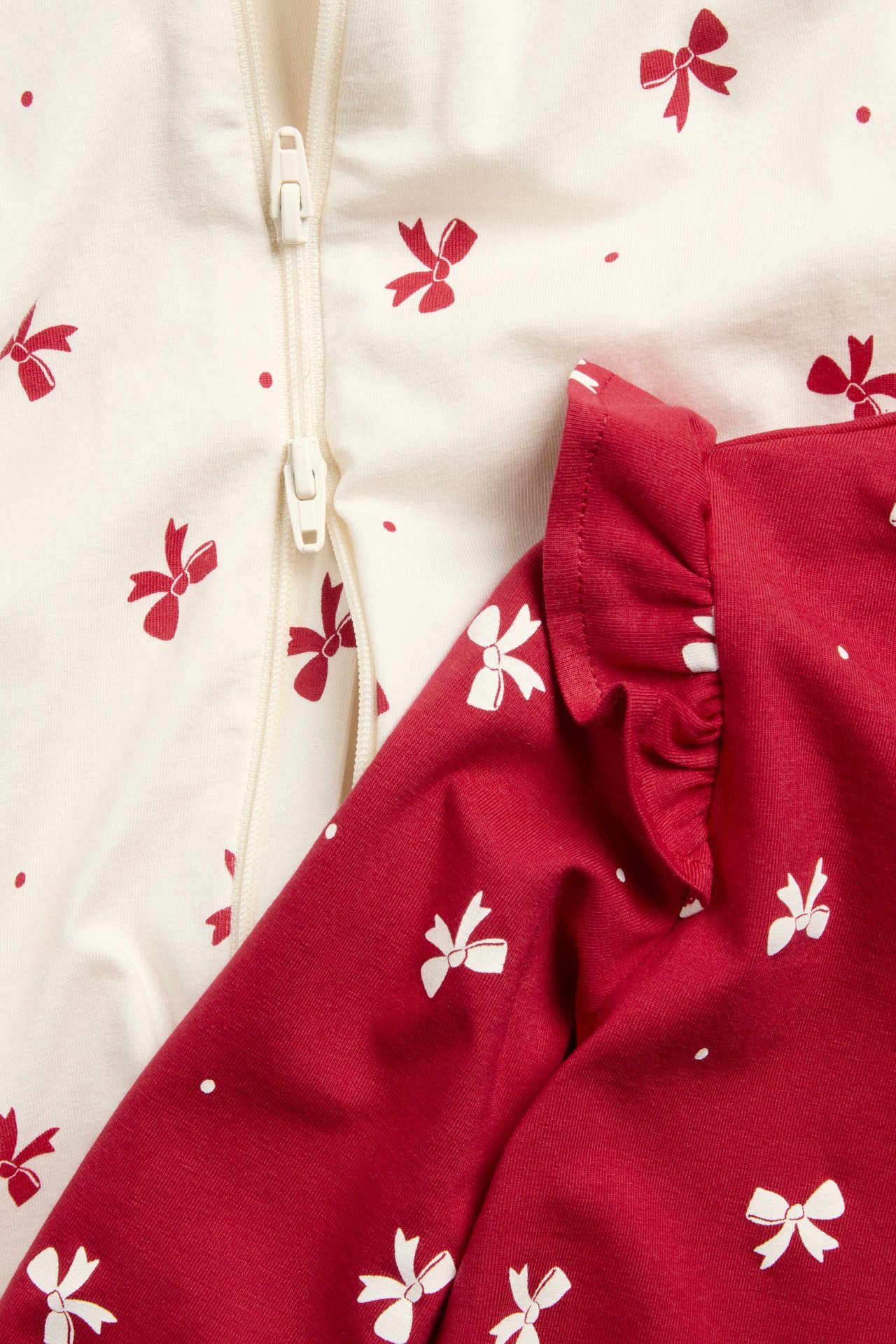 Zbliżenie na piżamy dla niemowląt 2-pak: kremowa z czerwonymi kokardkami i zamkiem, czerwona z białymi kokardkami i falbanką.