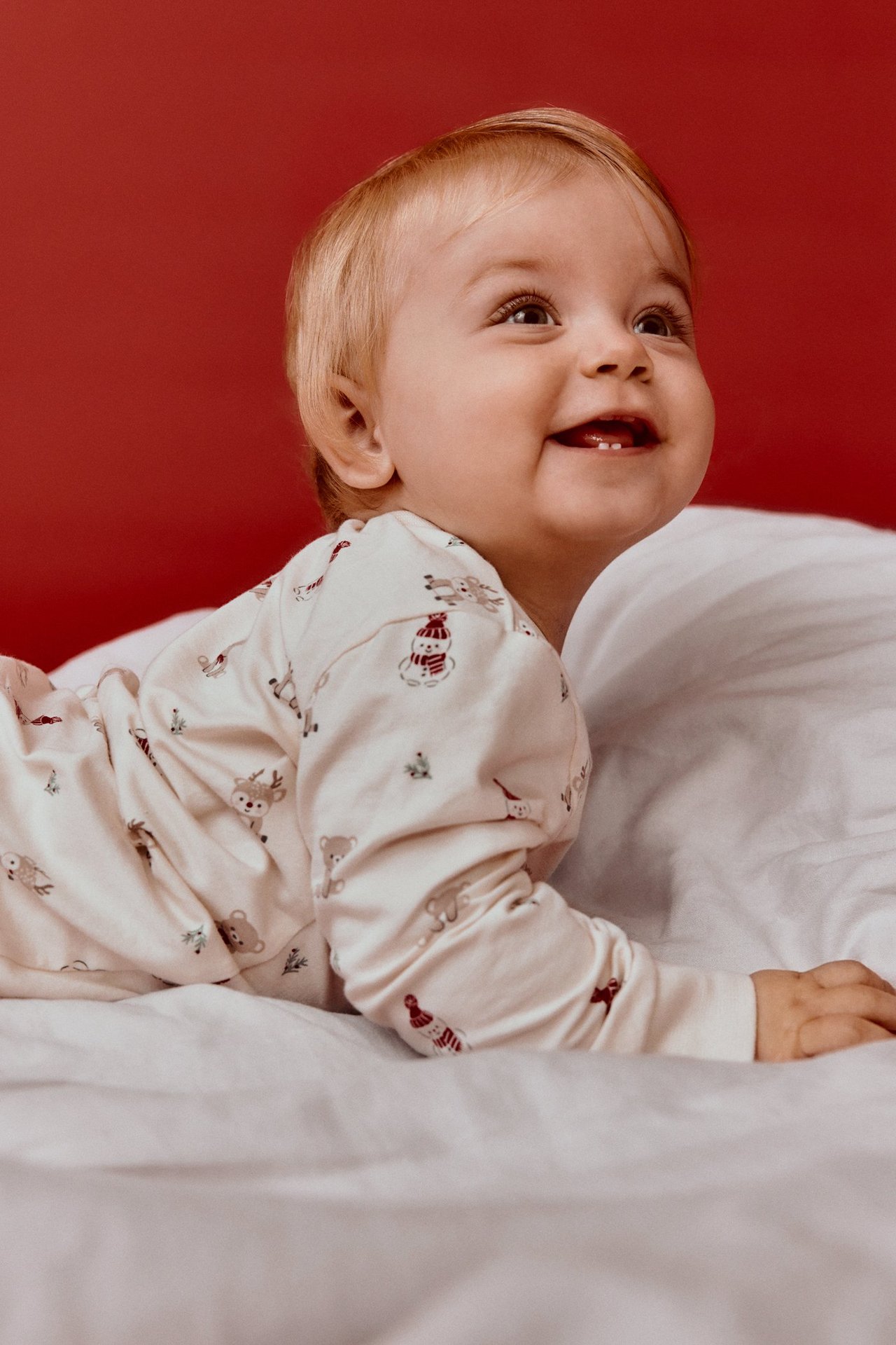 Lähikuva hymyilevästä vauvasta yllään luonnonvalkoiset kuviolliset joulupyjamat, maaten valkoisella alustalla.