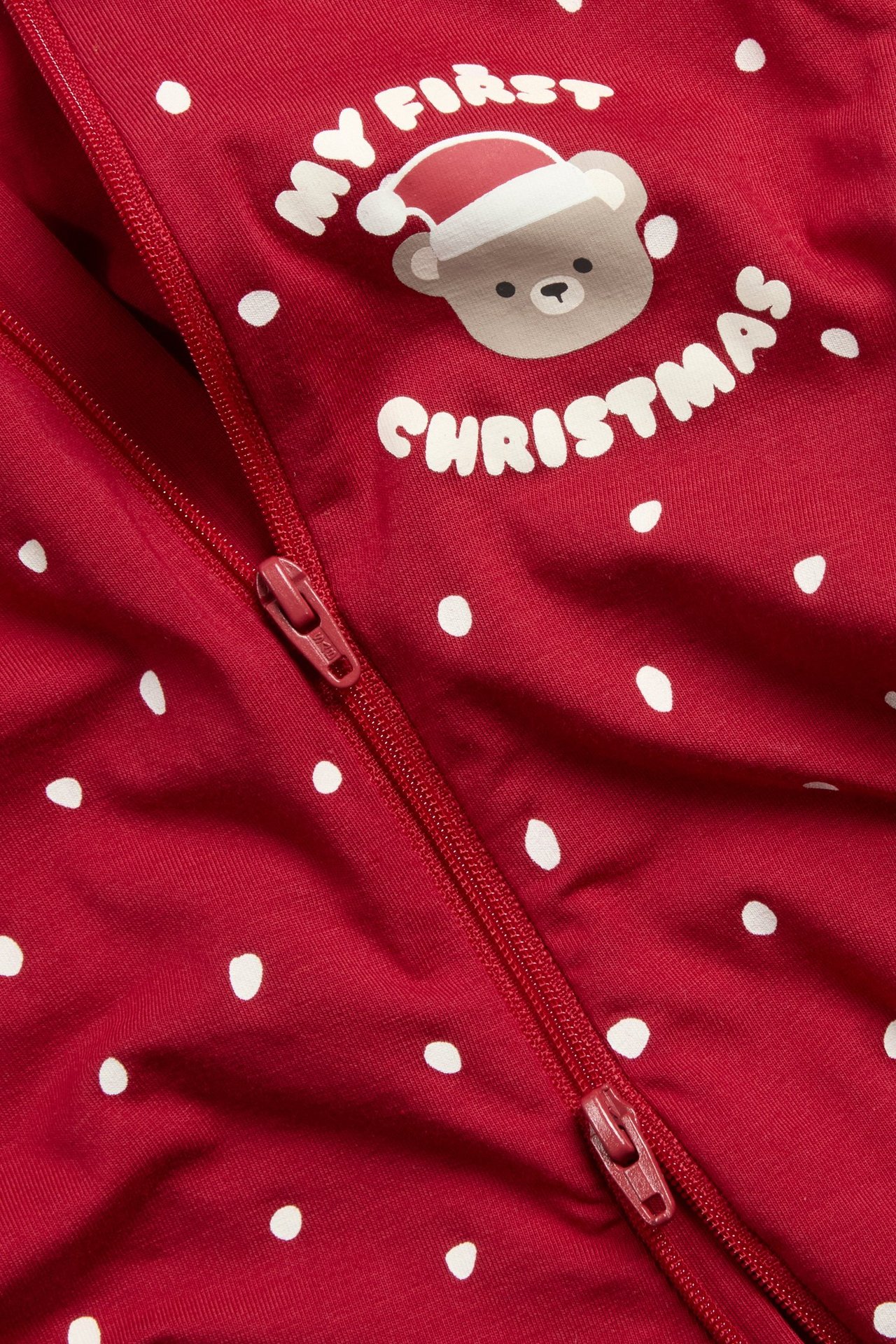 Lähikuva punaisesta vauvanpotkupuvusta, jossa valkoisia pilkkuja, kaksisuuntainen vetoketju ja 'My First Christmas' nallekuvio.